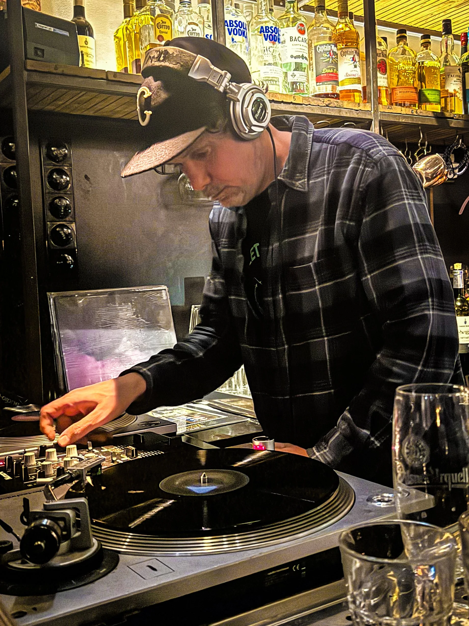 A man wearing headphones and a plaid shirt DJing at a bar, with liquor bottles on shelves behind him.