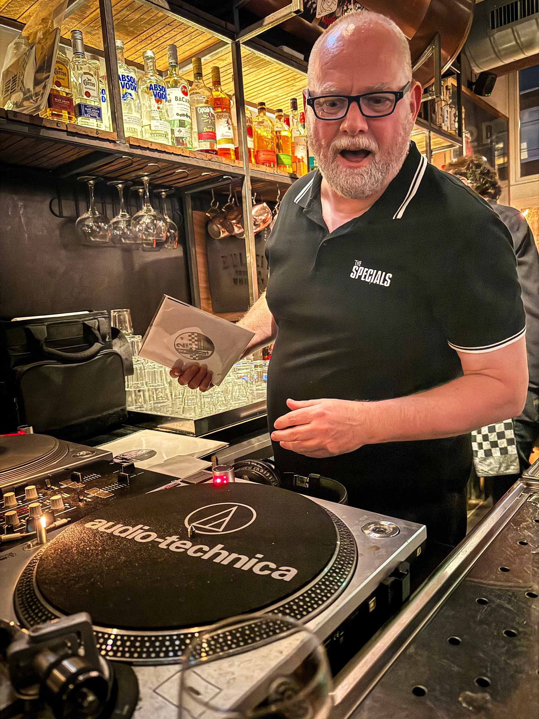 A man with glasses and a beard standing behind DJ equipment with a Pioneer turntable, liquor bottles on shelves, and wine glasses hanging upside down in a bar or nightclub setting.