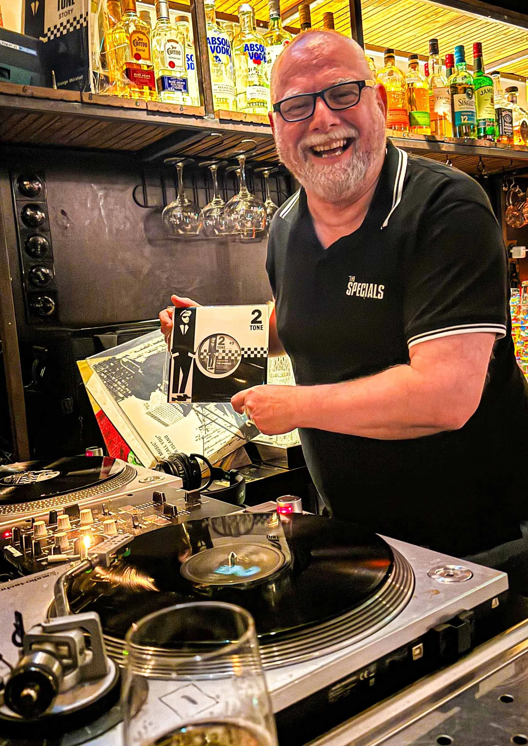 A man with glasses and a beard, smiling and laughing, standing behind DJ equipment at a bar or nightclub. Shelves in the background hold various bottles of alcohol, and glasses are hanging upside down. The man is holding a vinyl record case and appears to be in a lively environment.