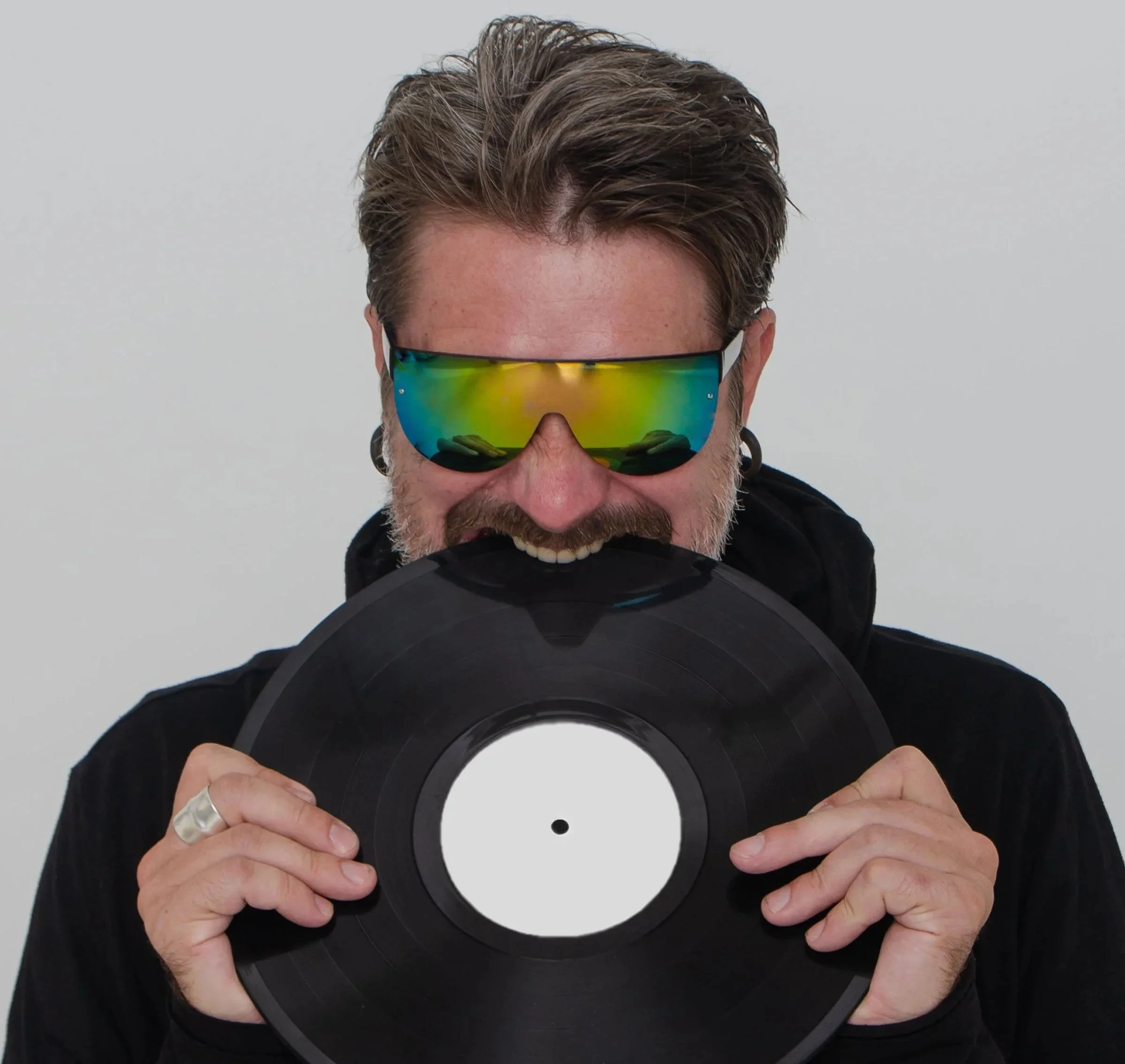 A portrait of DJ Necto Lucas - a man with a beard wearing mirrored sunglasses and a black jacket, holding a black vinyl record in front of his face, against a plain light gray background.