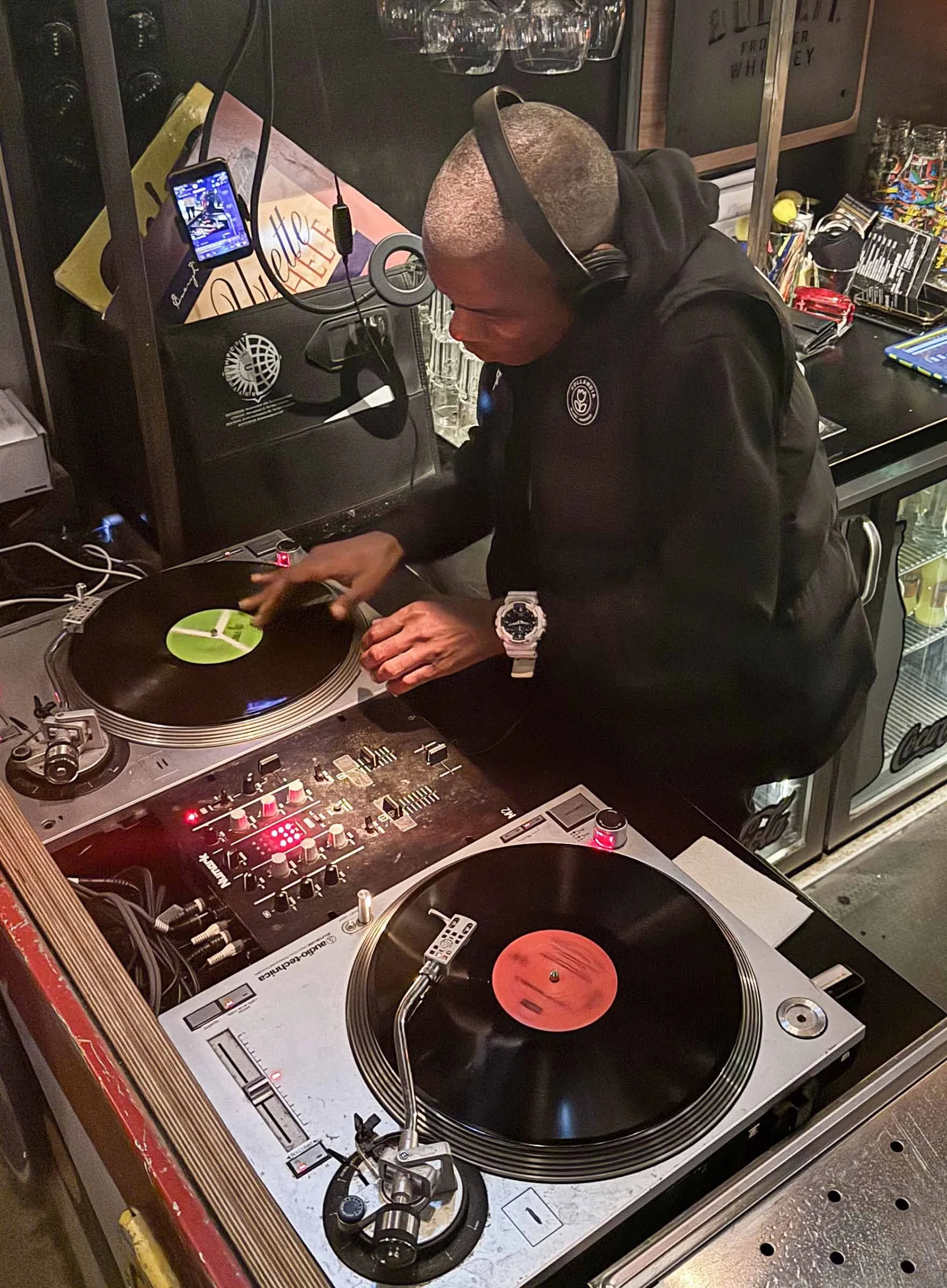 A man with a shaved head and headphones is DJing with turntables and a mixer. The scene is at a bar or club with drinks, media equipment, and a beverage refrigerator in the background.
