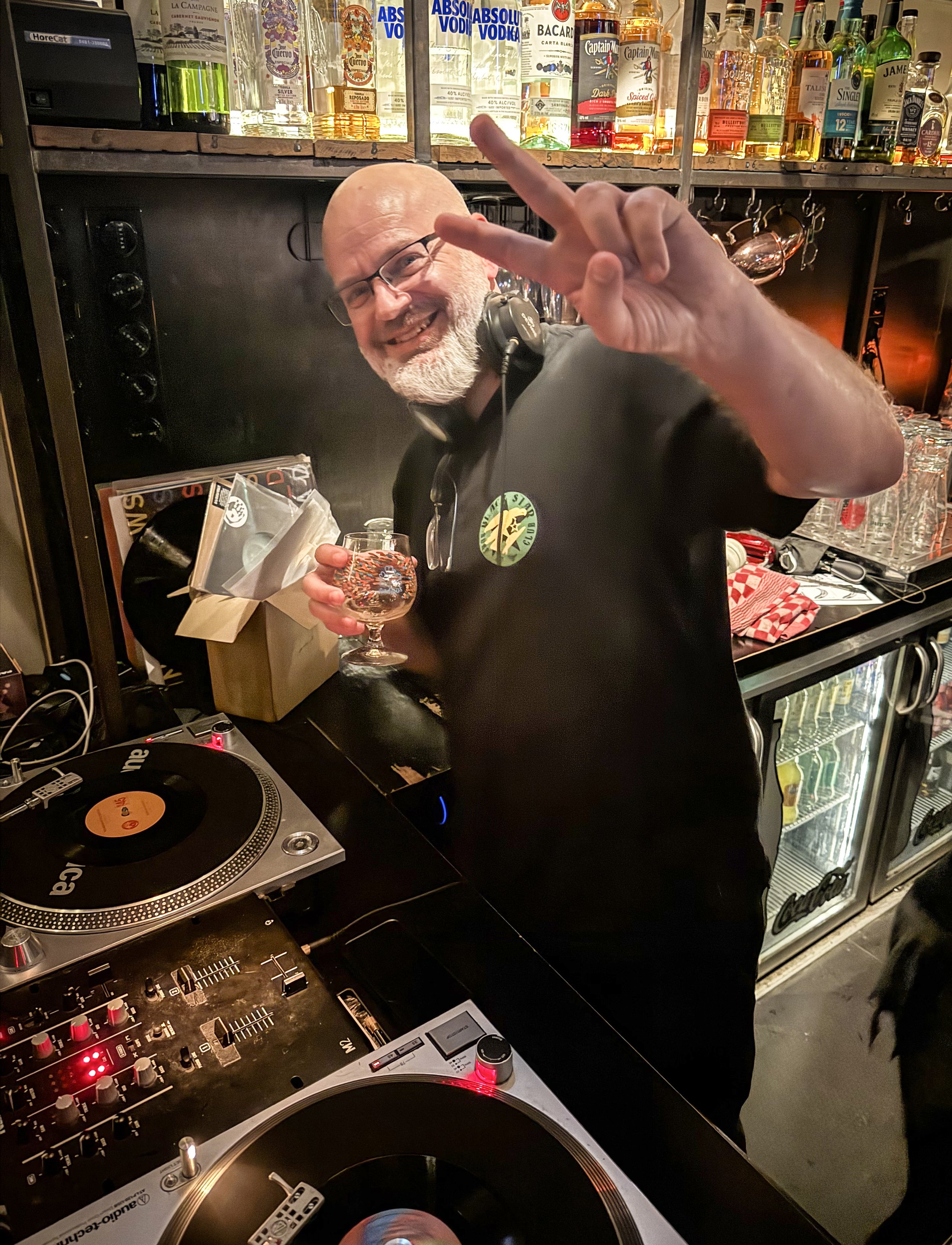 A man with glasses and a beard smiling and holding a glass of drink, making a peace sign with his fingers, standing behind a DJ mixer in a bar, with liquor bottles on shelves behind him.