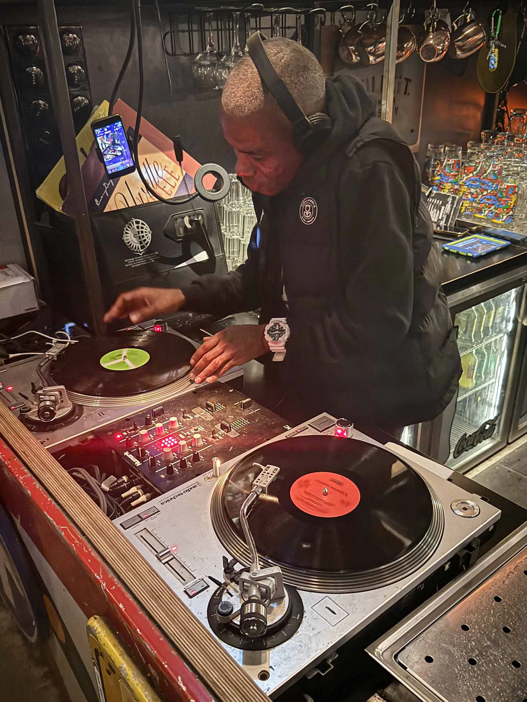 A DJ wearing headphones, a black hoodie, and a large watch, using turntables and a mixer in a bar or club setting with glasses and hanging copper pots in the background.
