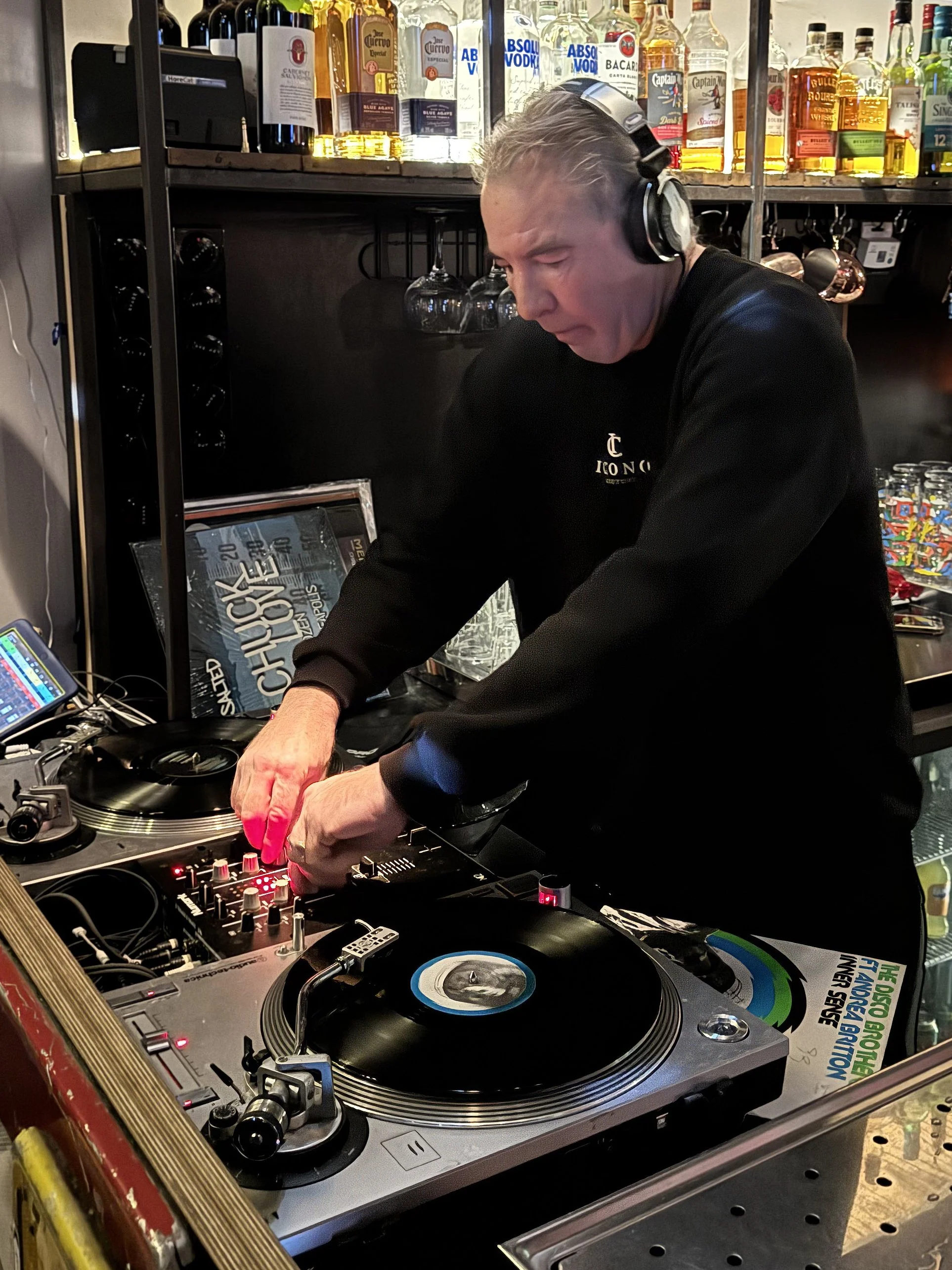 A DJ wearing headphones operates turntables and mixer in a bar or club, with liquor bottles displayed on shelves behind him.