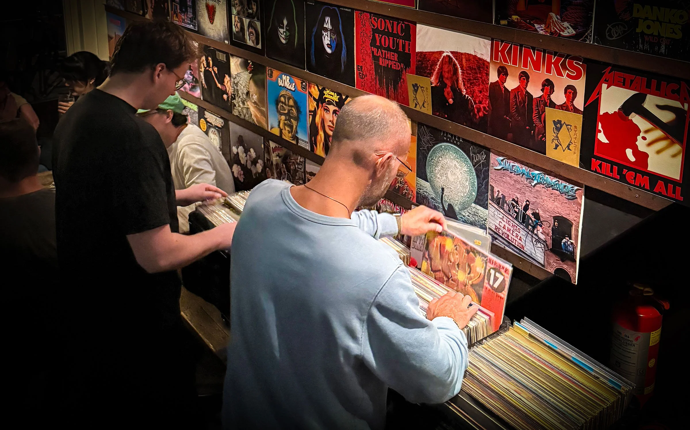 People browsing vinyl records at an Amsterdam Vinyl Club live event. There are album covers displayed on shelves, including bands like Metallica, Kinks, and Sonic Youth.