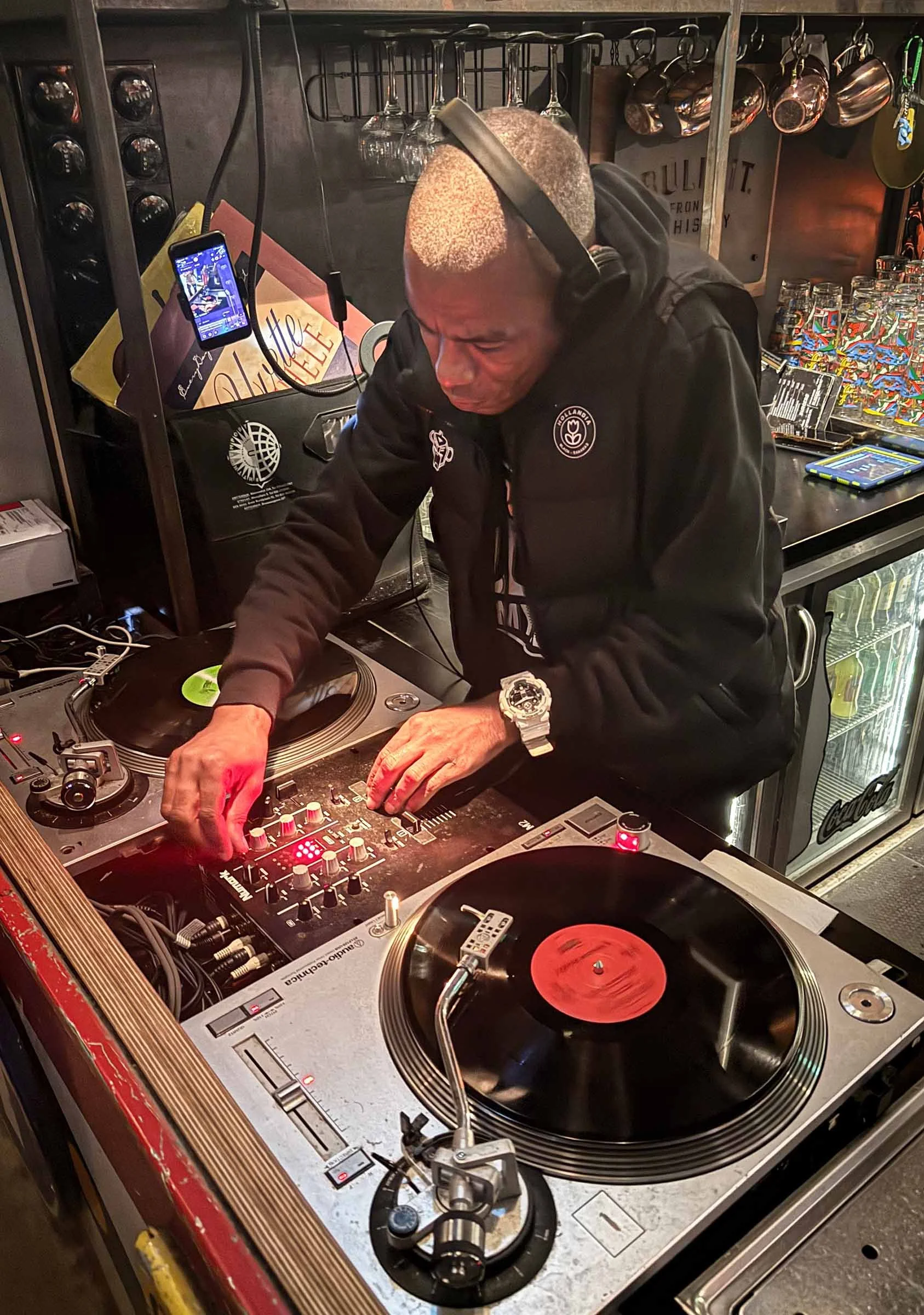 A DJ with a closely shaved head wearing a black hoodie with a logo, headphones, and a large silver watch, is mixing music on a DJ turntable and mixer in a bar or club setting, with glassware and kitchen utensils hanging in the background.