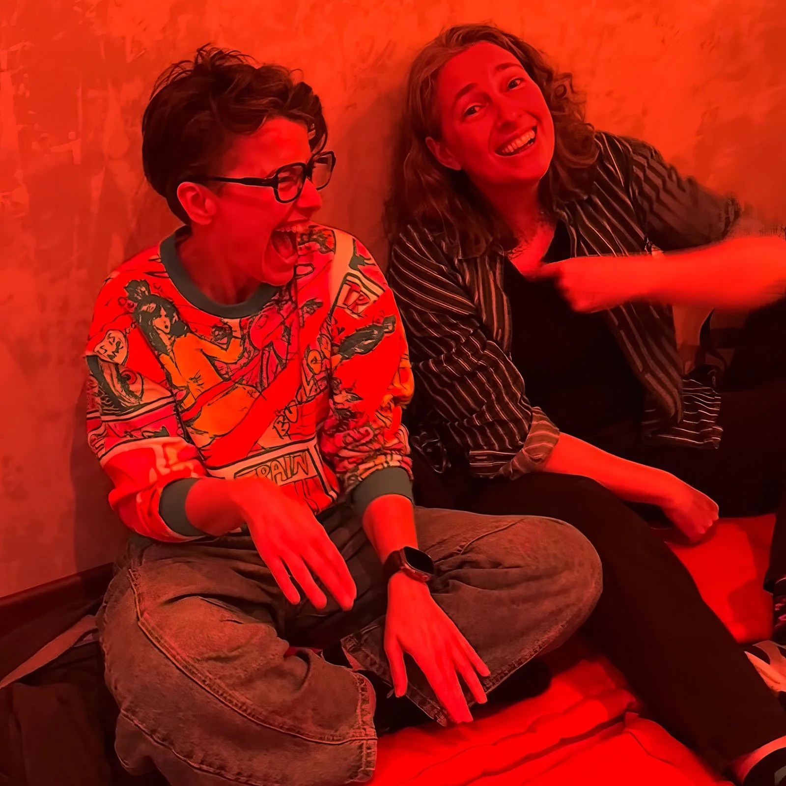 Two people having fun at an Amsterdam Vinyl Club live event. The person on the left is wearing glasses and a colorful graphic sweatshirt, laughing with their mouth open. The person on the right has curly hair and is smiling, wearing a striped shirt.
