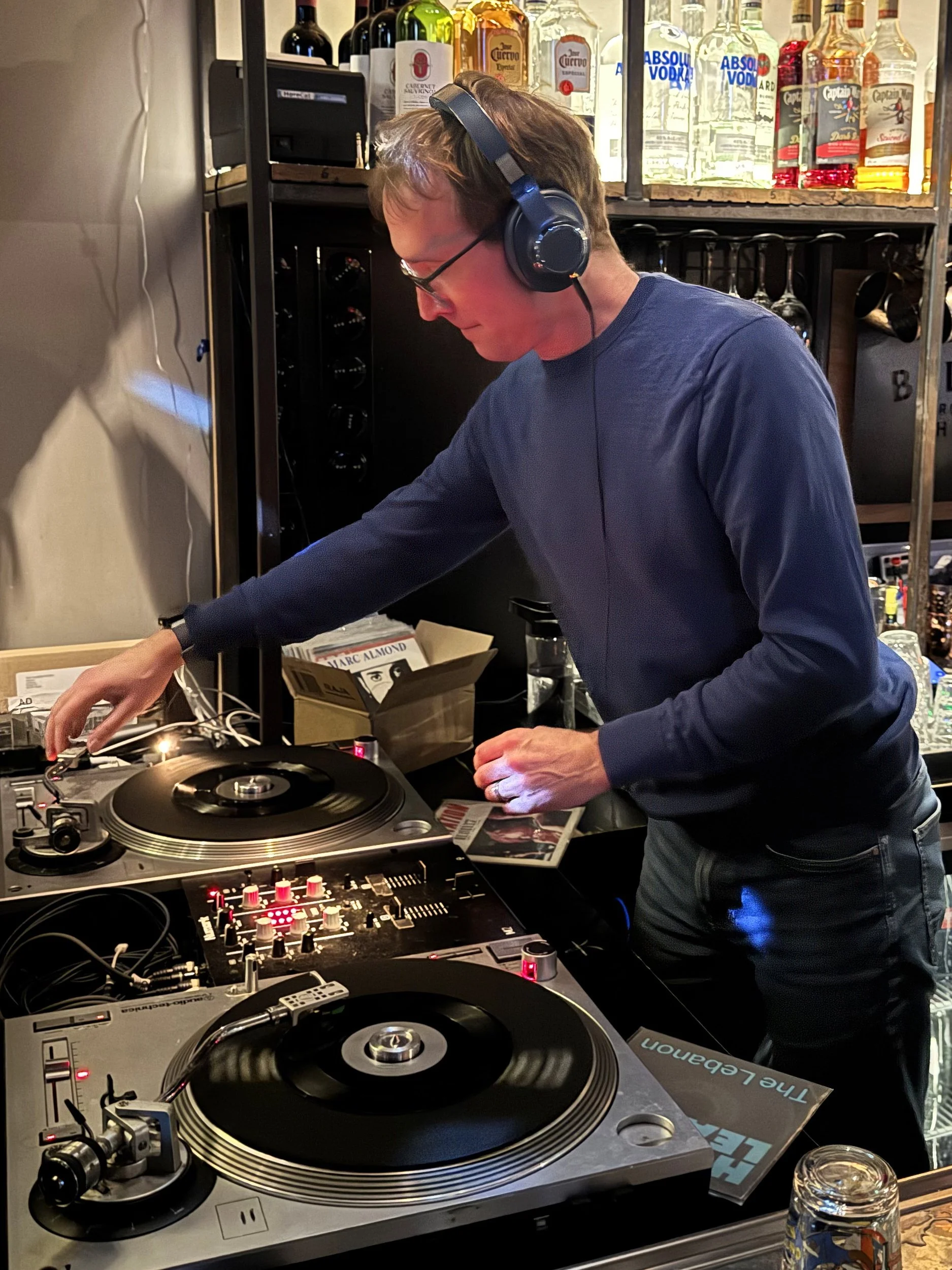 A man in a blue shirt and glasses DJing with headphones, using turntables and a mixer in a bar with bottles of alcohol on shelves in the background.