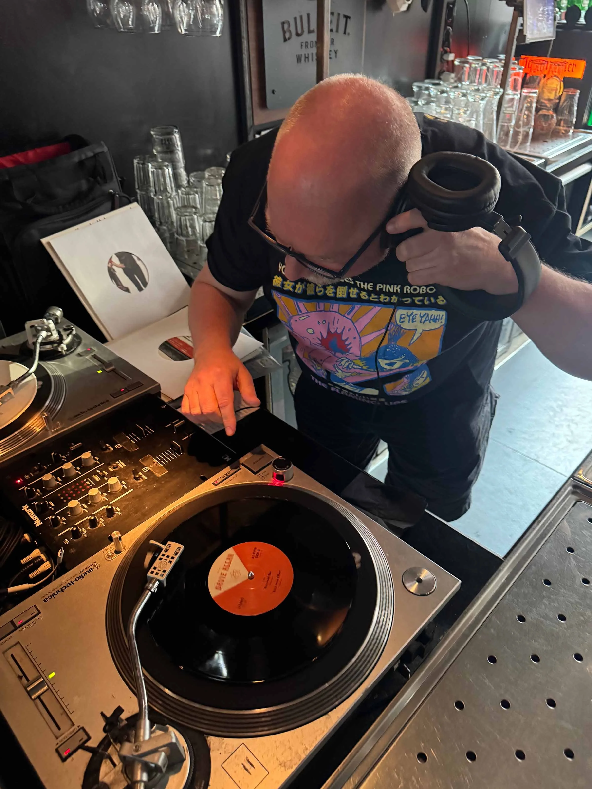 A person with a bald head and glasses DJing at a turntable with vinyl records, wearing headphones, in a bar or music venue with glassware and bottles in the background.