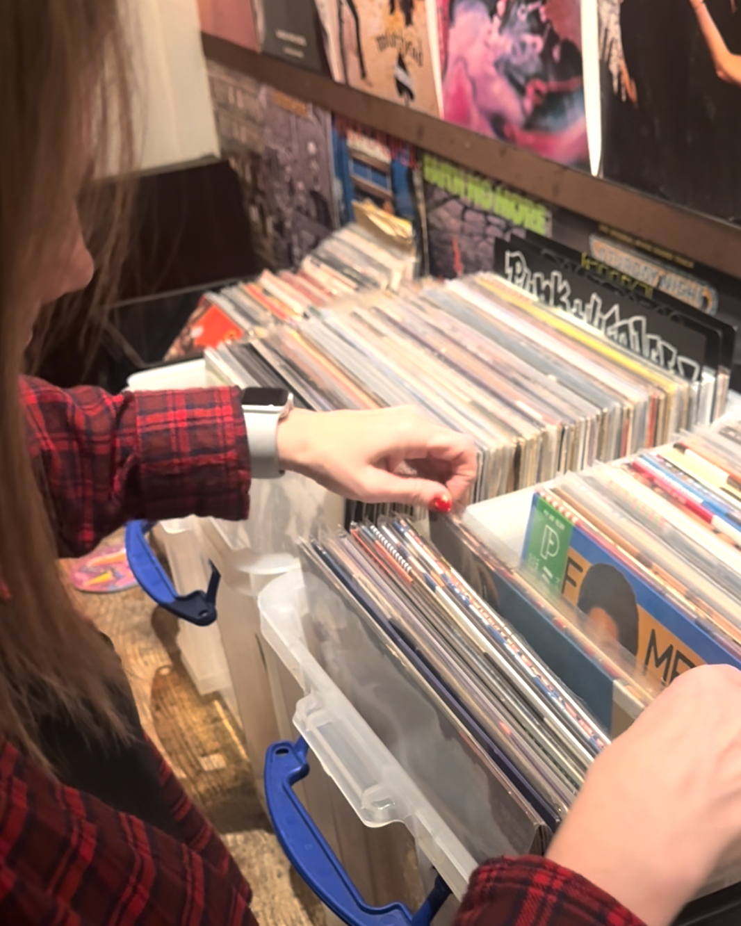 A person with long hair wearing a red plaid shirt and a smart watch is browsing through vinyl records at Amsterdam Vinyl Club.