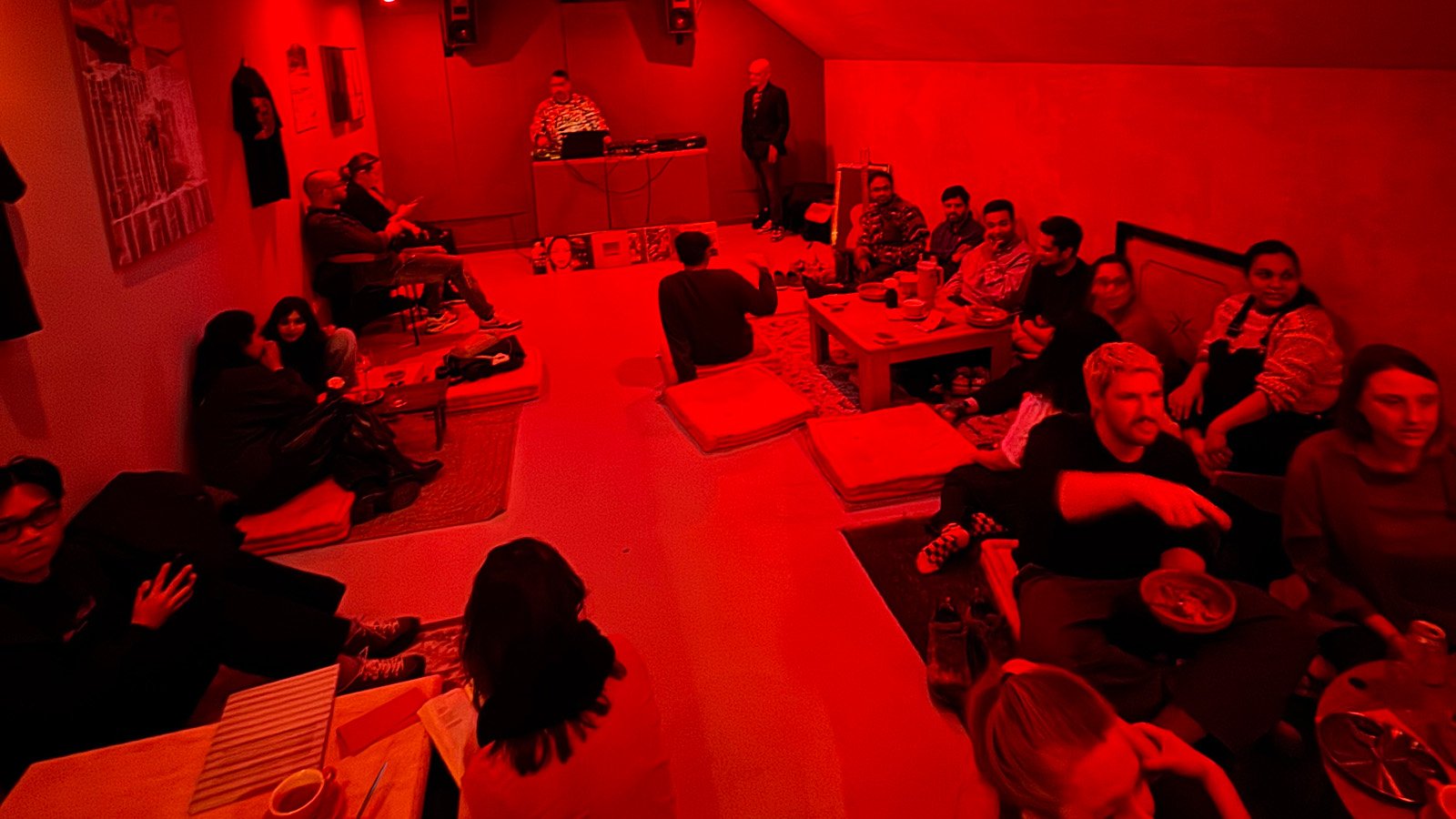 An Amsterdam Vinyl Club monthly event in a city venue - people gathered in a dimly lit room with red lighting, sitting around tables, some eating and socializing.