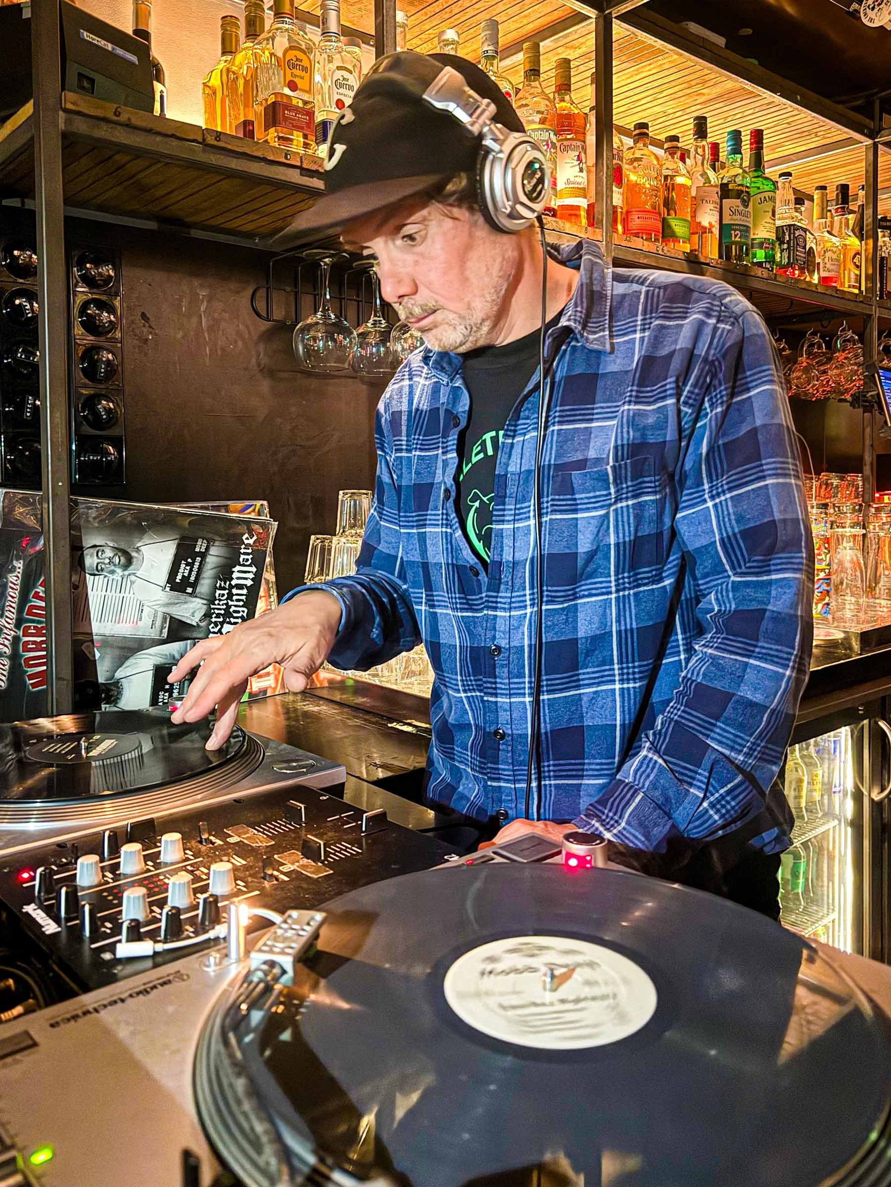 A DJ wearing headphones and a baseball cap is mixing music on DJ equipment in a bar or nightclub setting. There are bottles of liquor and glasses arranged on shelves in the background.