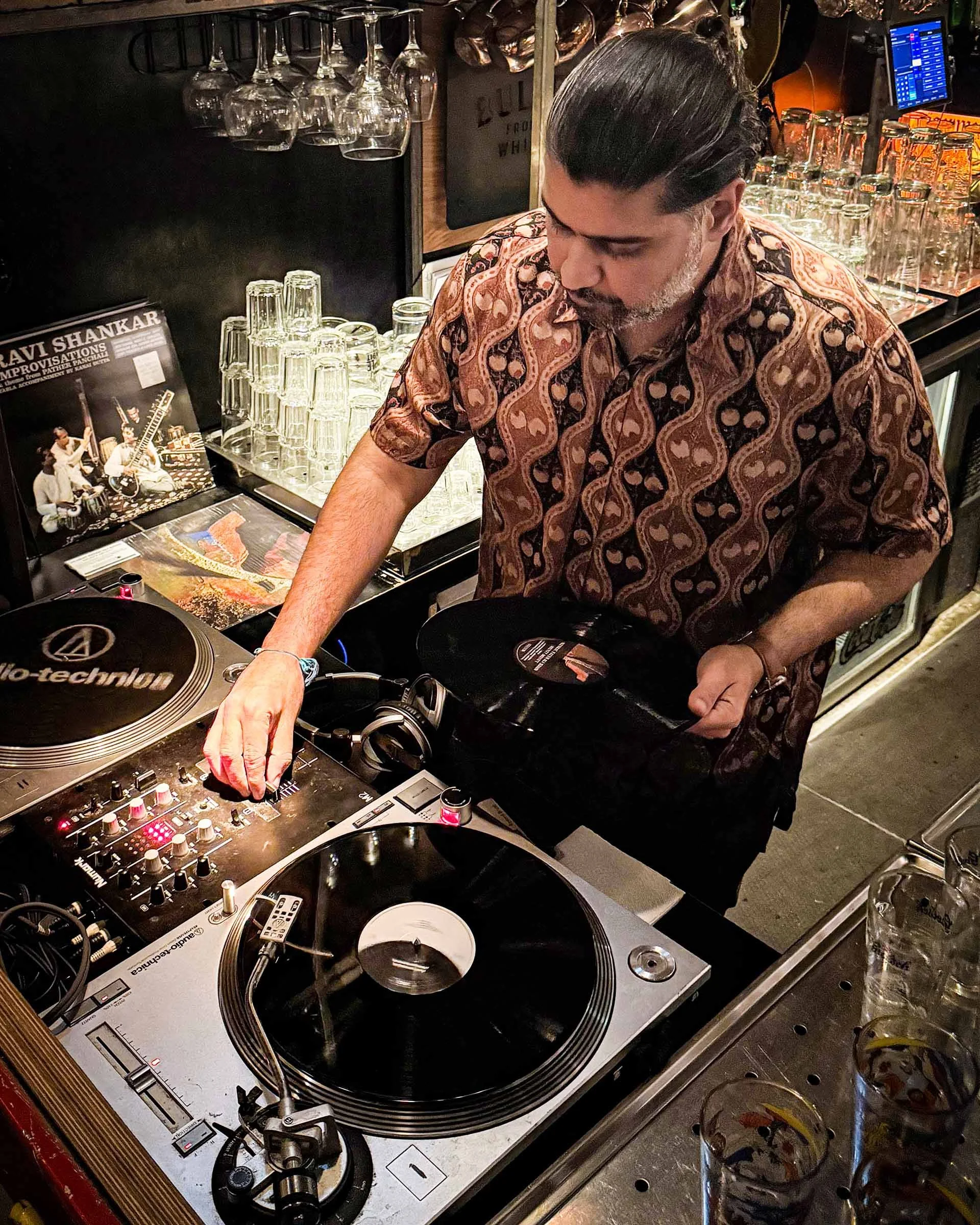 A DJ wearing a patterned shirt is standing behind DJ equipment, including turntables and a mixer, in a bar setting with glasses and bottles in the background.