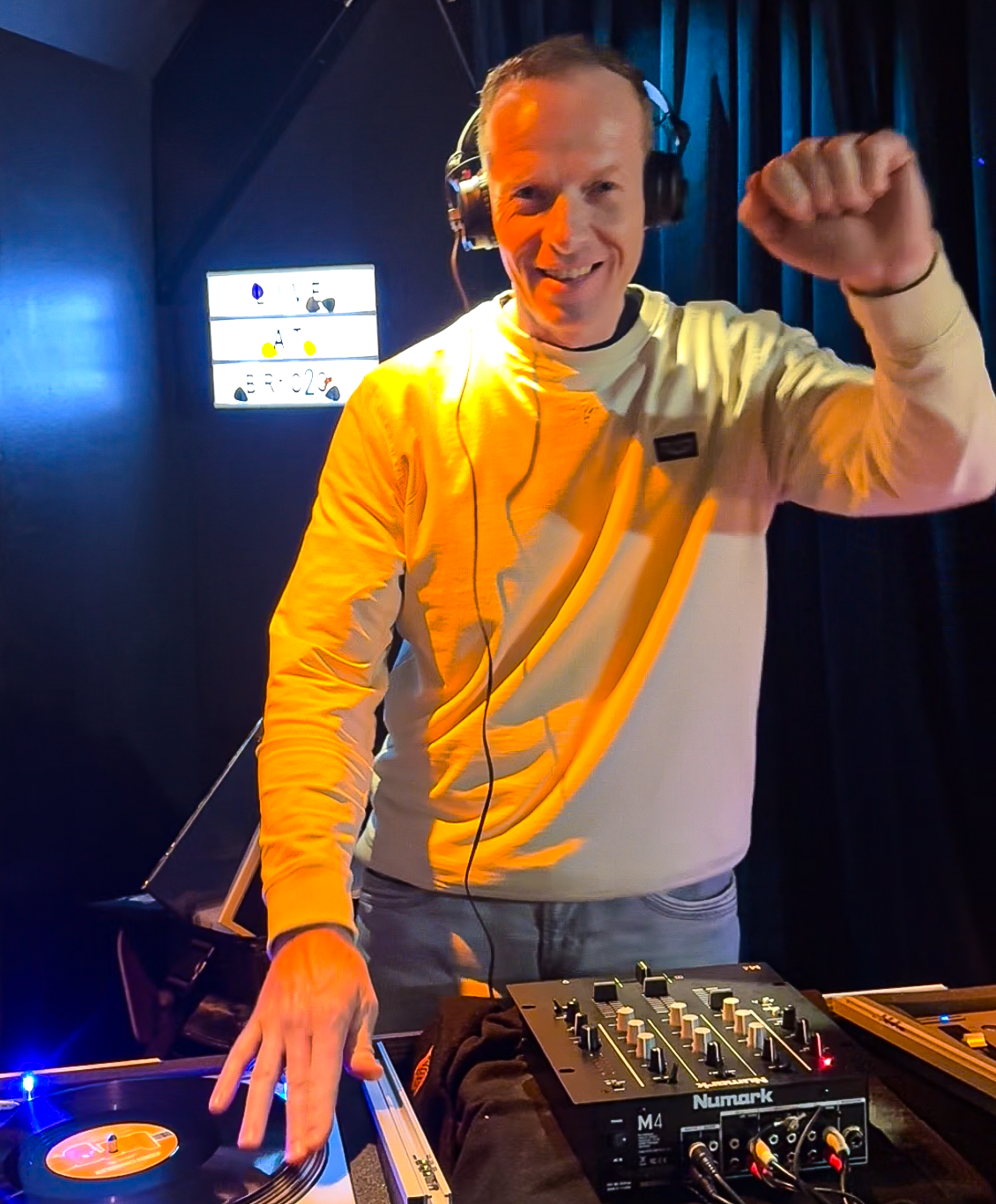 A smiling man wearing headphones and a yellow sweater, standing behind DJ equipment, raising his right fist in celebration, with a dark background and a bright display screen in the background.