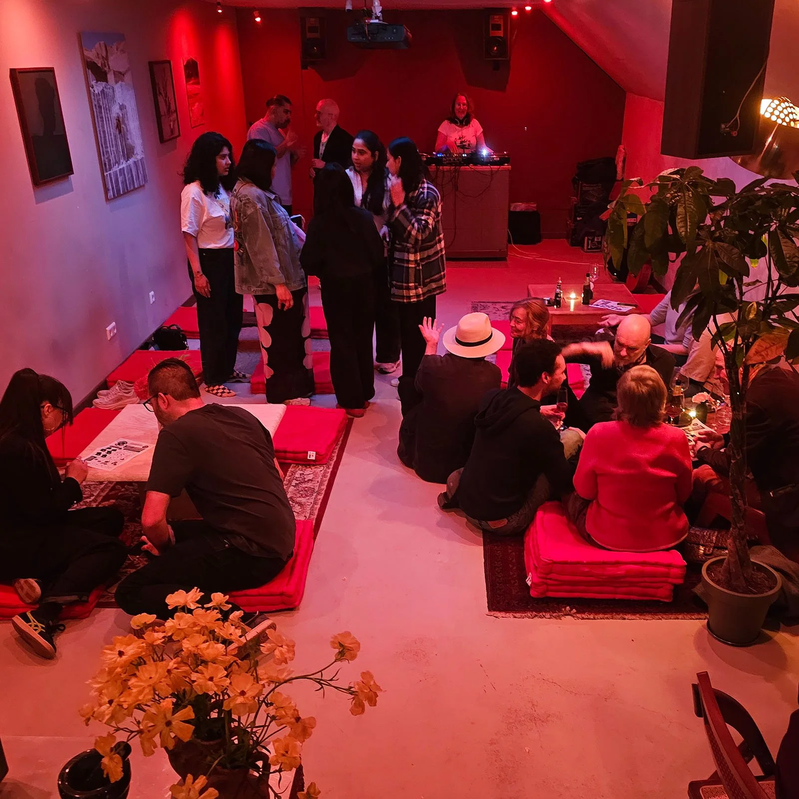 A lively indoor Amsterdam Vinyl Club event, with people engaging in conversations. Some are sitting on cushioned floor seats, while others are standing and chatting. A DJ is playing music in the background, creating an energetic atmosphere.