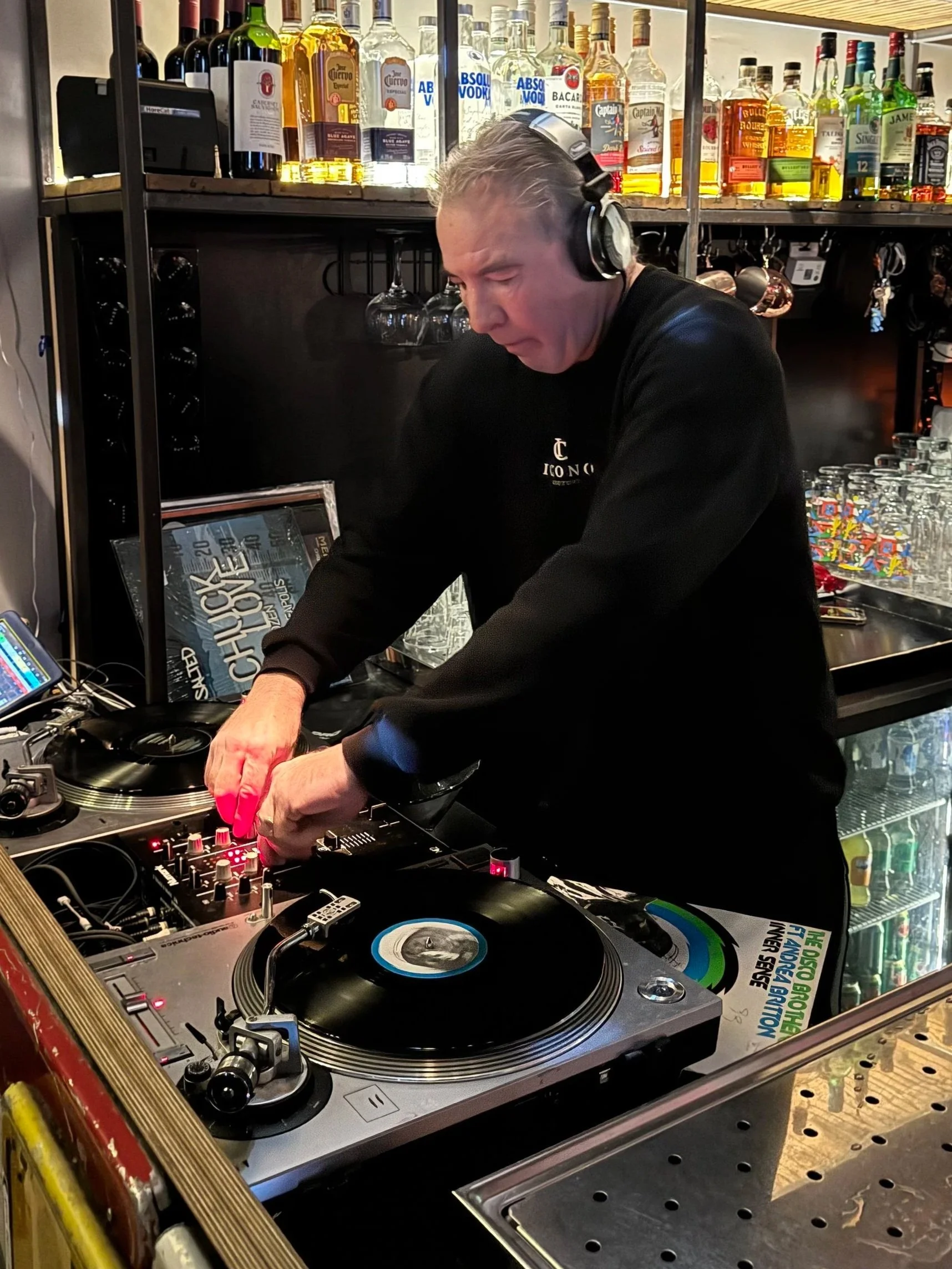 A DJ wearing headphones is playing music behind a mixer and turntables in a bar with bottles of alcohol on shelves in the background.