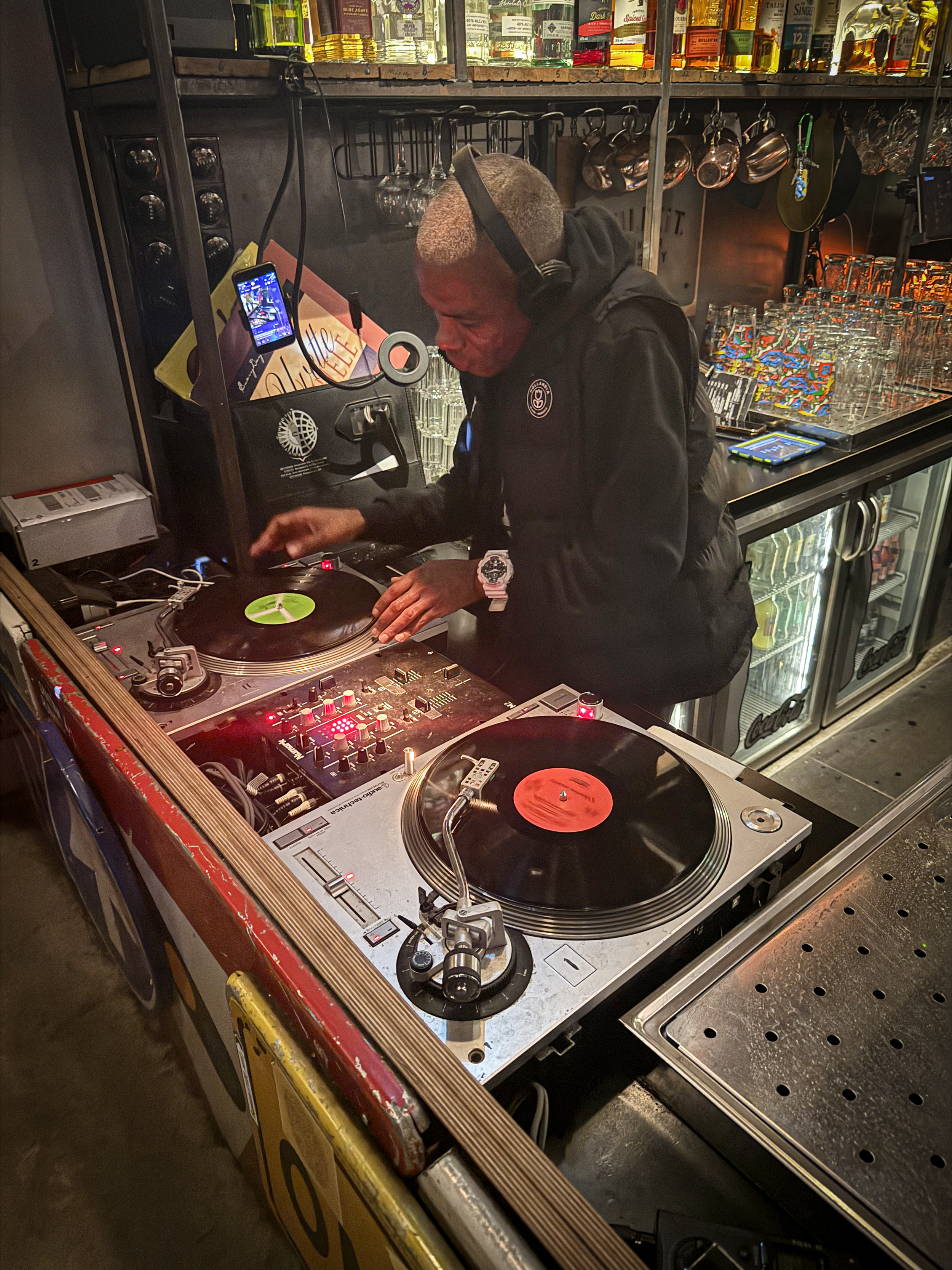 A DJ is mixing records on a turntable in a bar or club. He is wearing a black jacket, a large white watch, and headphones. Shelves behind him hold bottles of alcohol, glasses, and cups.