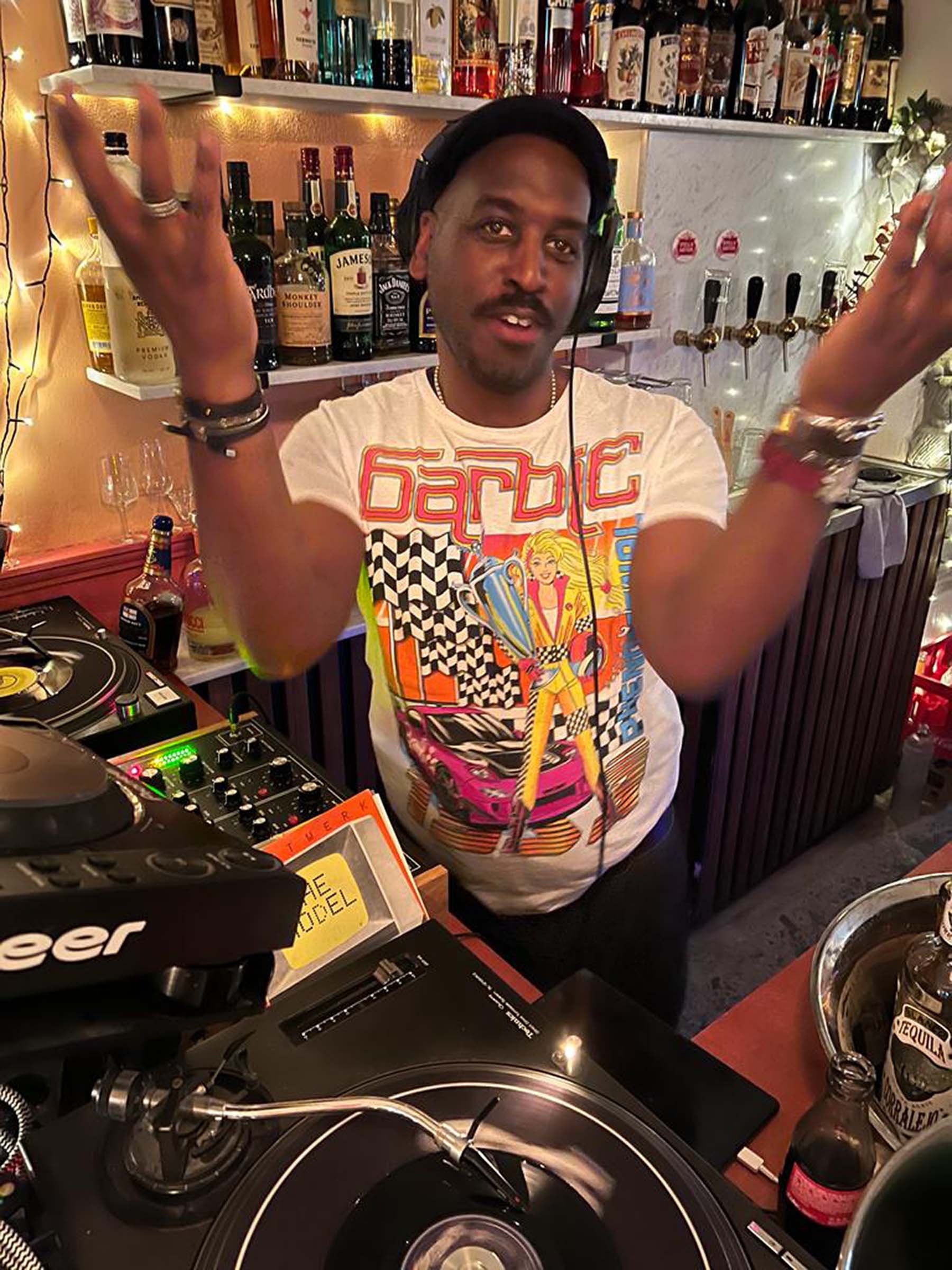 A man wearing headphones and a colorful cartoon graphic t-shirt standing behind DJ equipment in a bar setting with shelves of liquor bottles and string lights in the background.