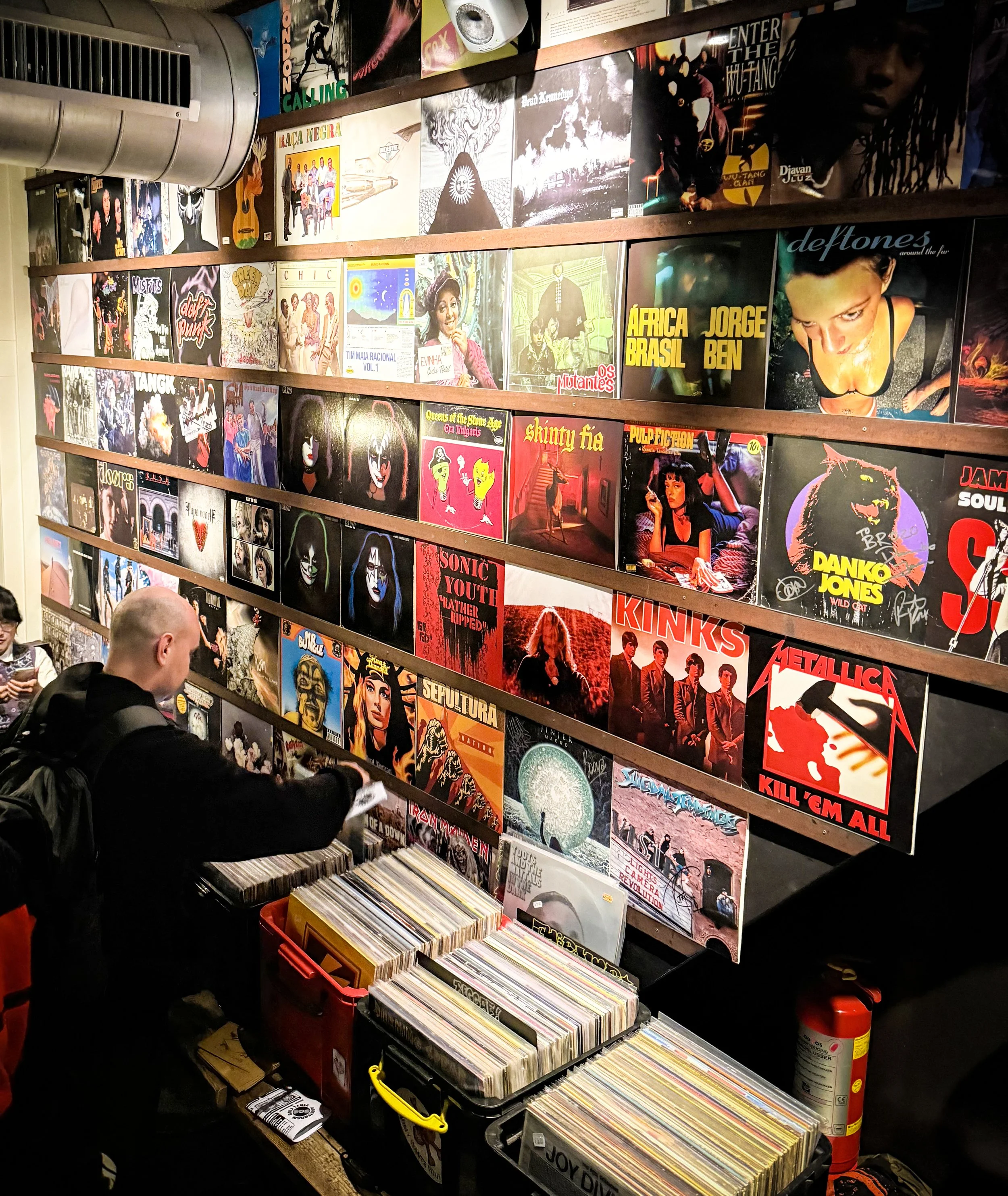 A person shopping for vinyl records at a store with shelves of album covers on the wall, including artists like Metallica, Kinks, Sepultura, and Deftones.