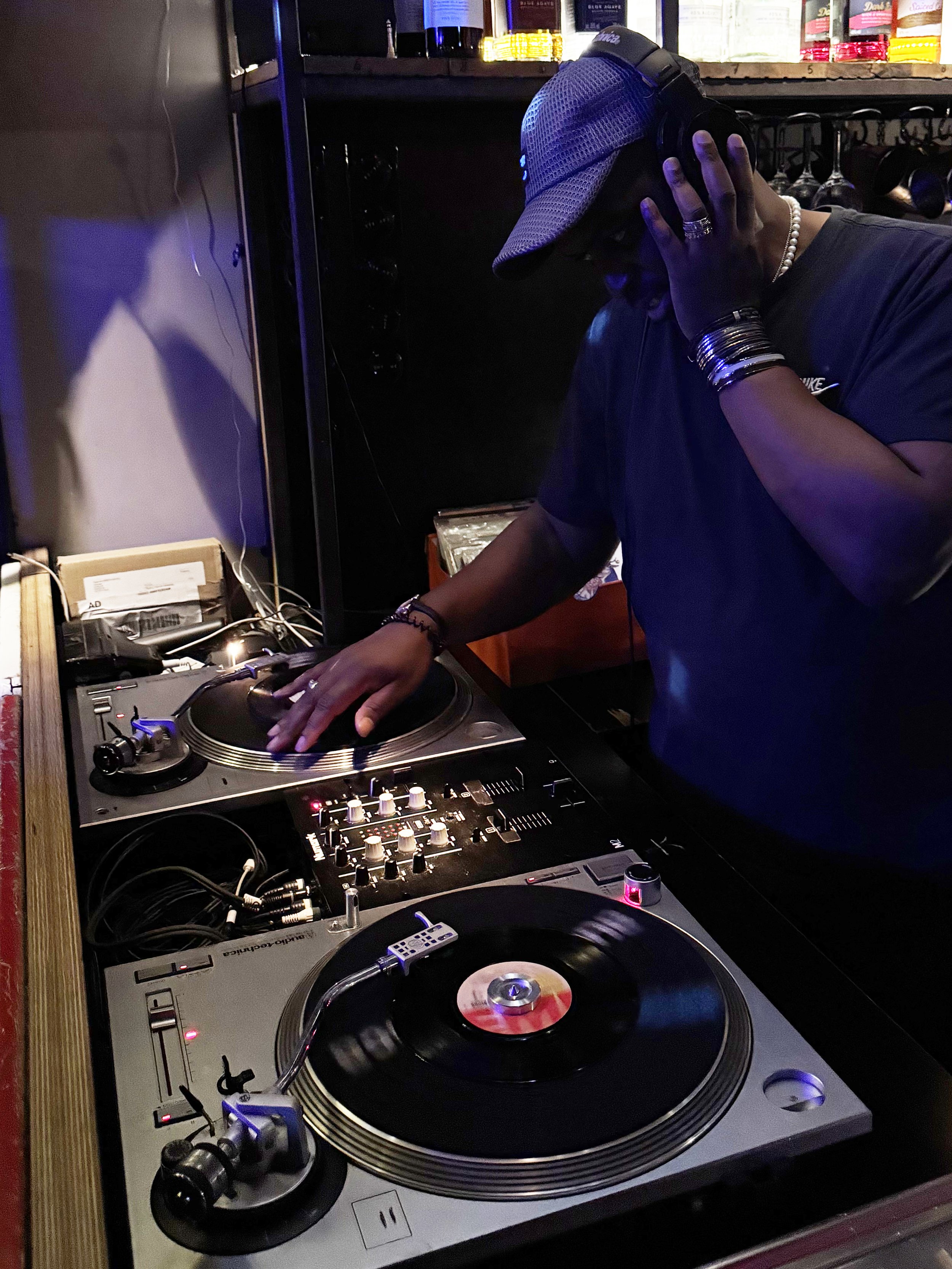 A DJ wearing a cap and jewelry using turntables and mixer in a dimly lit setting, possibly a bar or club.