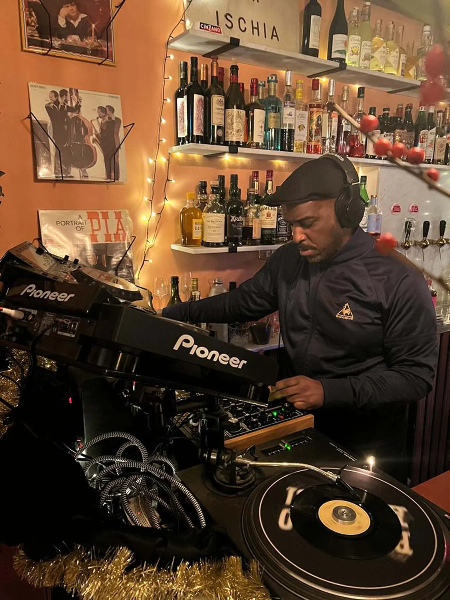 DJ wearing headphones operating a Pioneer DJ controller on a decorated bar counter with alcohol bottles on shelves and Christmas decorations in the background.