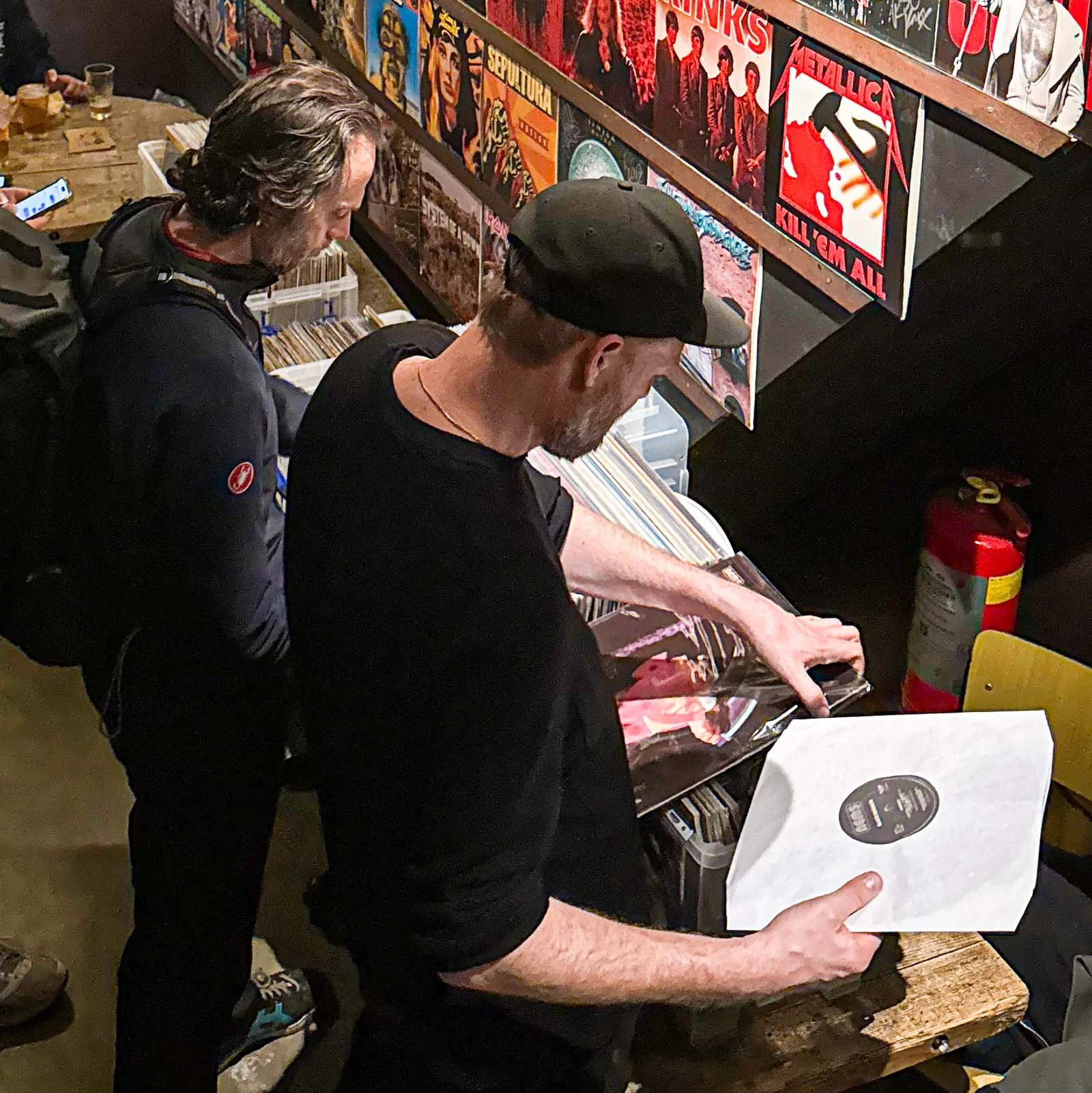 Two men are browsing vinyl records at an Amsterdam Vinyl Club live event. One is holding a record album, and the other is looking at records on the shelf. The wall is decorated with various colorful album covers.