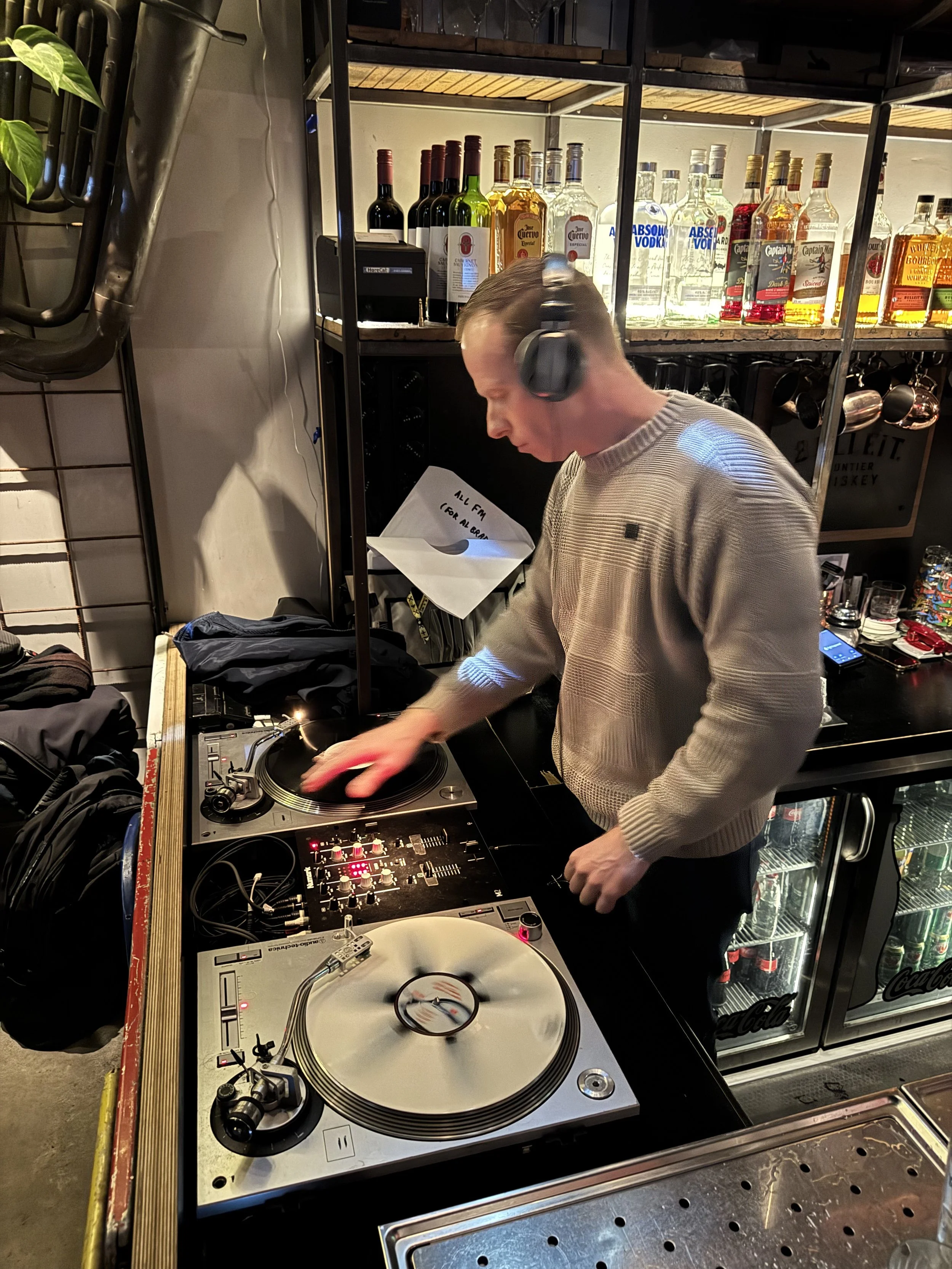 A man DJing with headphones on, using turntables and a mixer at a bar or club, with liquor bottles on shelves behind him.