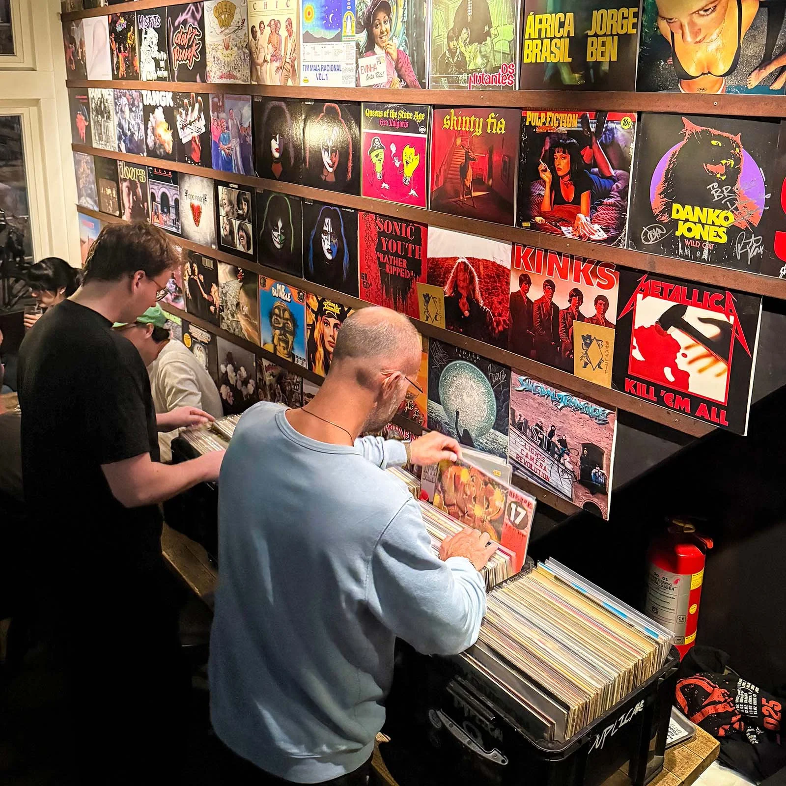 People browsing and selecting vinyl records at an Amsterdam Vinyl Club live event, with colorful album covers displayed on the wall behind them.