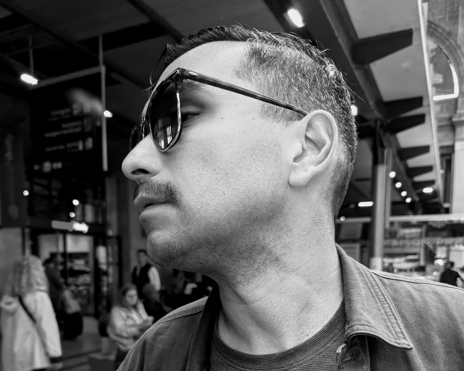 A black-and-white photo of a man wearing sunglasses looking to his left in a public indoor setting with people and shelves in the background.