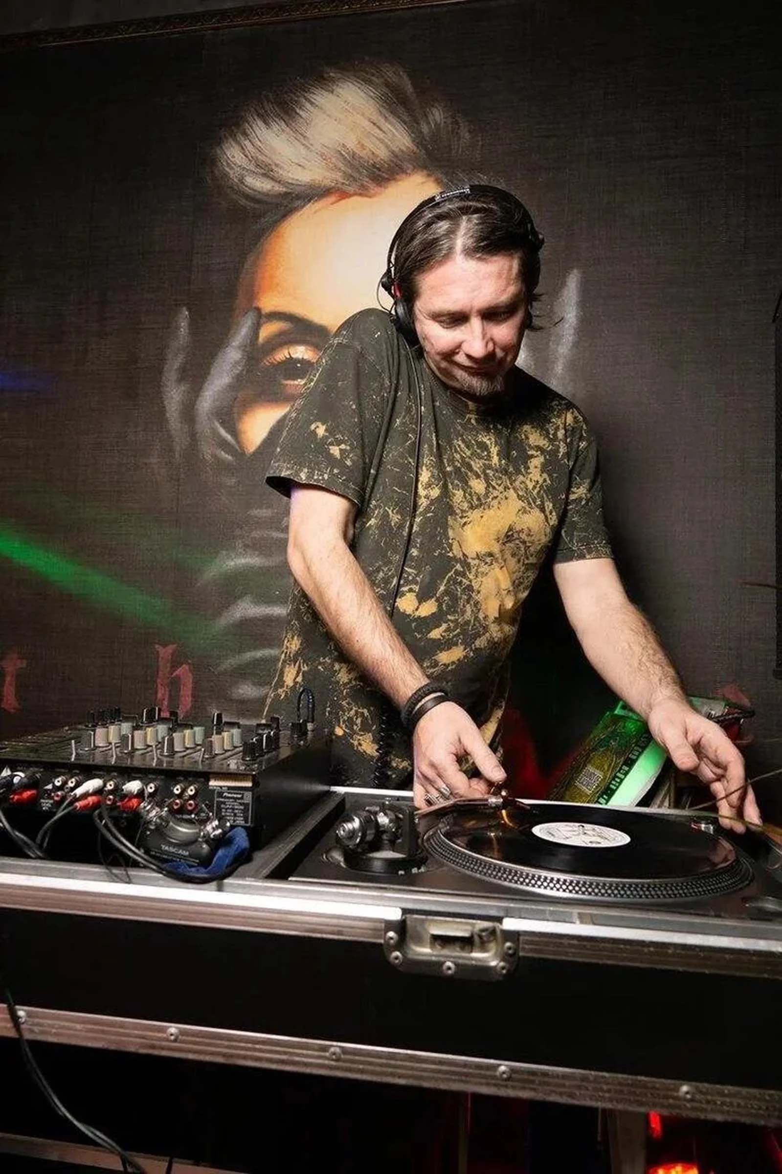 A portrait of DJ Necto Lucas wearing headphones and a black and gold t-shirt mixing music on a DJ controller at a club or event. Behind him, a large poster features a woman with styled hair and makeup.