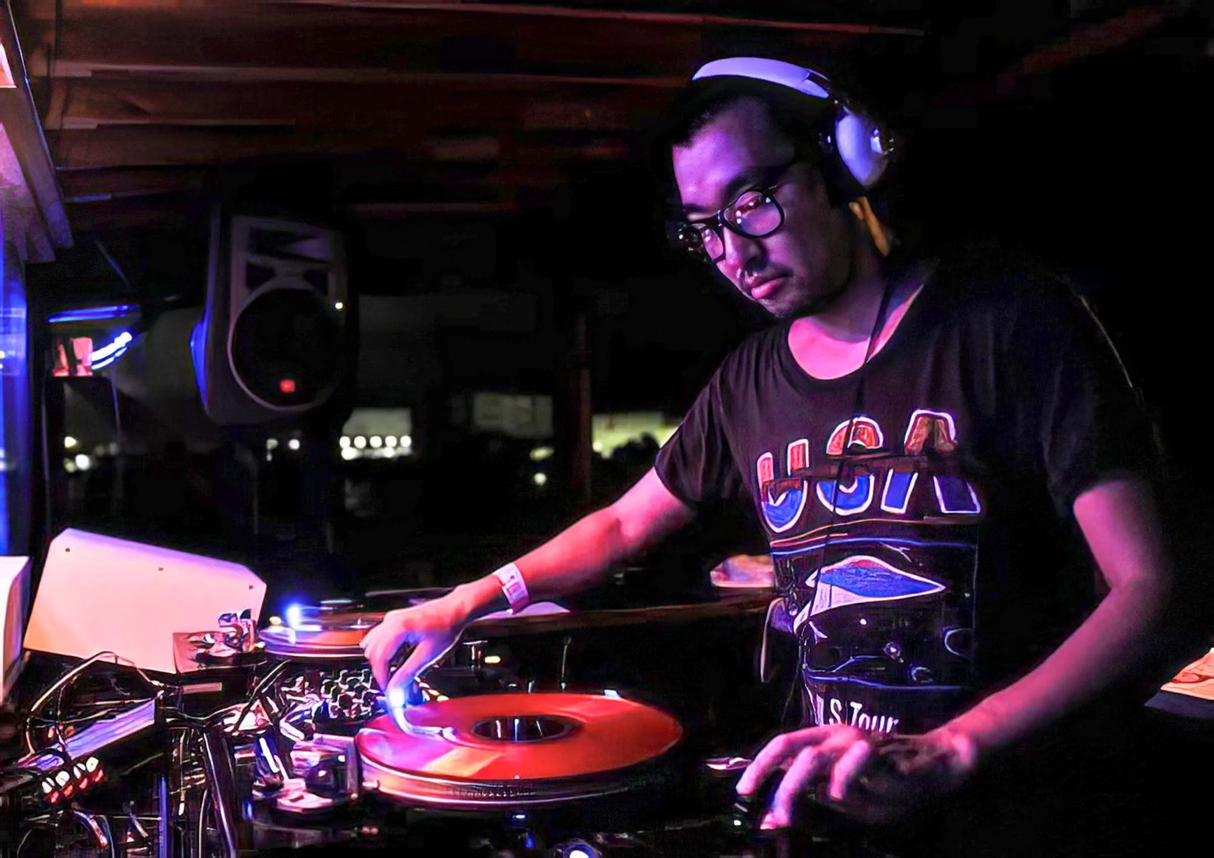 A young man with glasses and headphones DJing at a dark indoor event, illuminated by colorful lights.