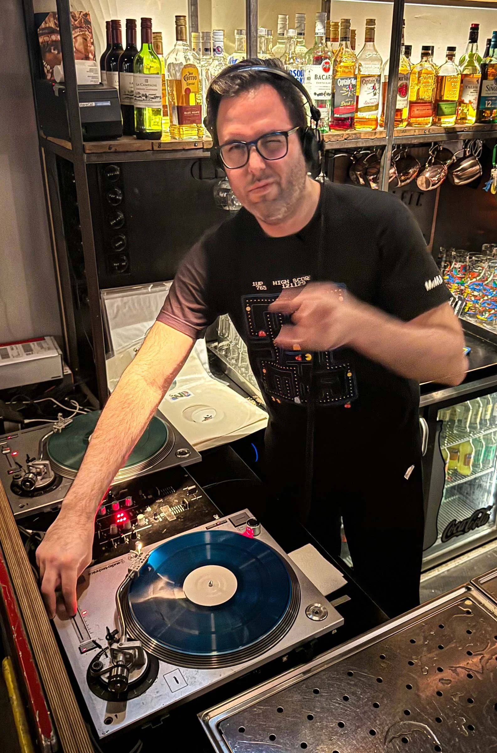 A man with glasses and headphones, DJing with a turntable and mixing equipment in a bar or restaurant with bottles of alcohol on shelves behind him.
