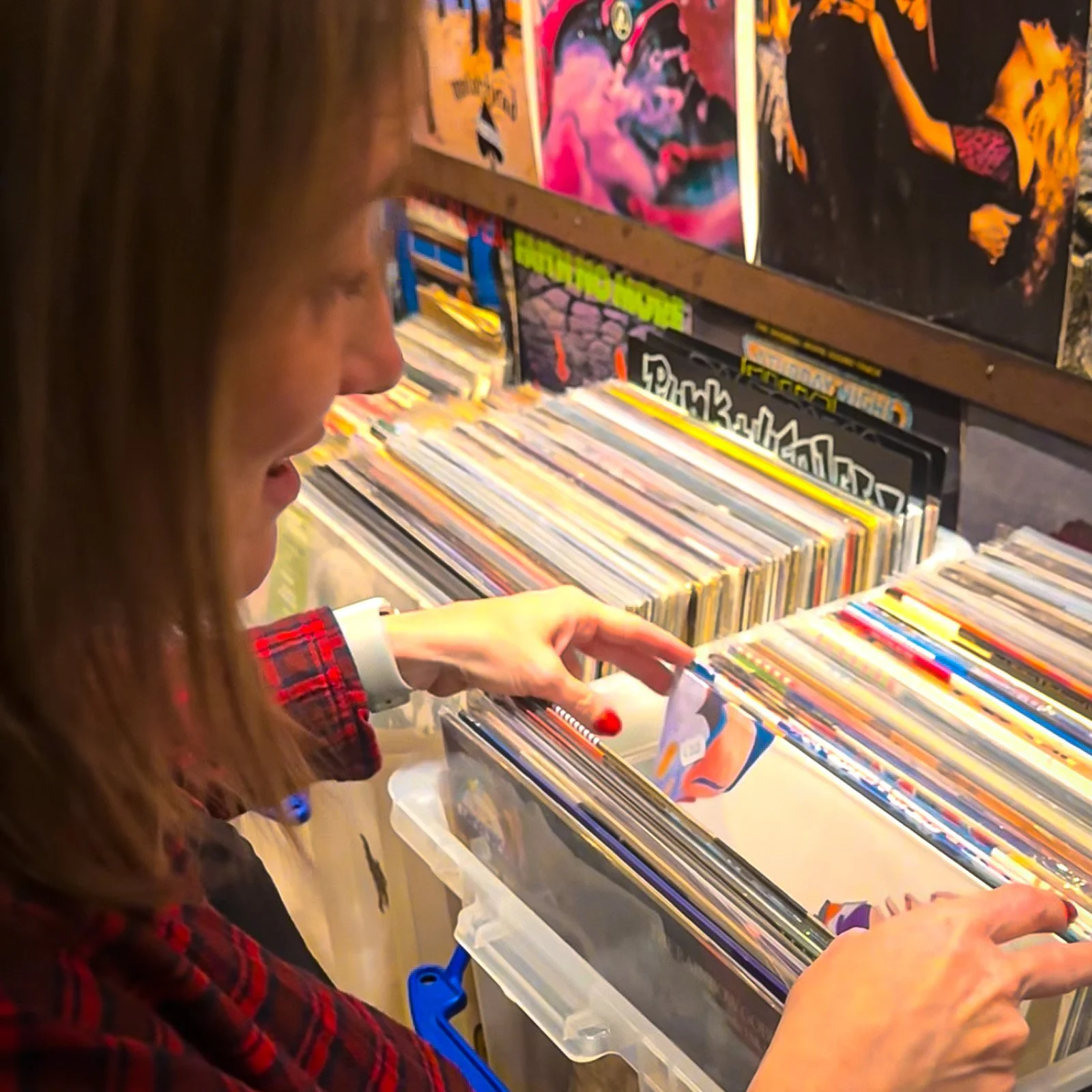 A woman browses through a collection of vinyl records at an Amsterdam Vinyl Club live event. She is wearing a red and black plaid shirt and examining a record in her hand. The shelves behind her are filled with colorful album covers.