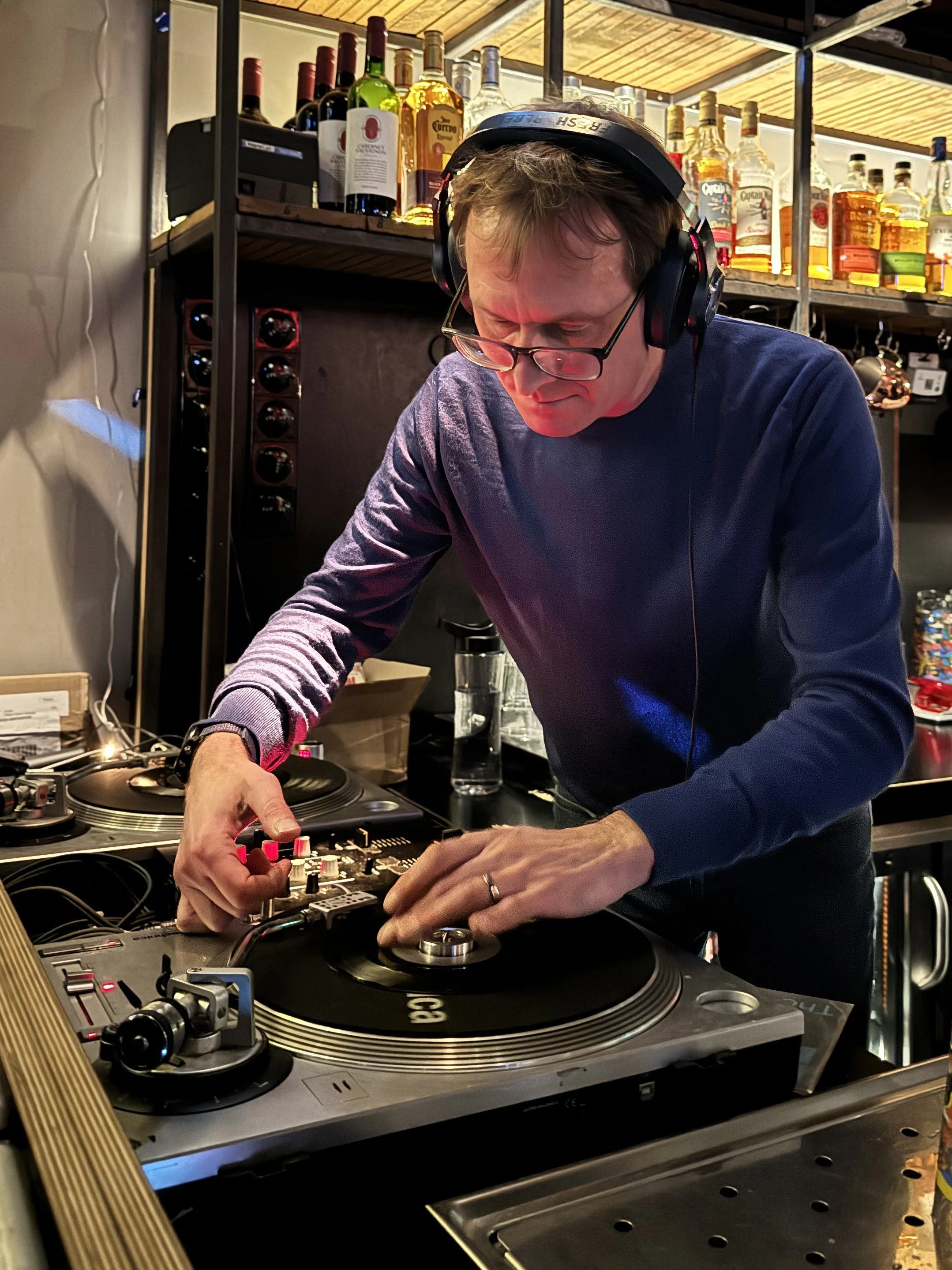 A DJ wearing glasses, black headphones, and a blue sweater is operating turntables in a bar or club setting, with liquor bottles on shelves behind him.