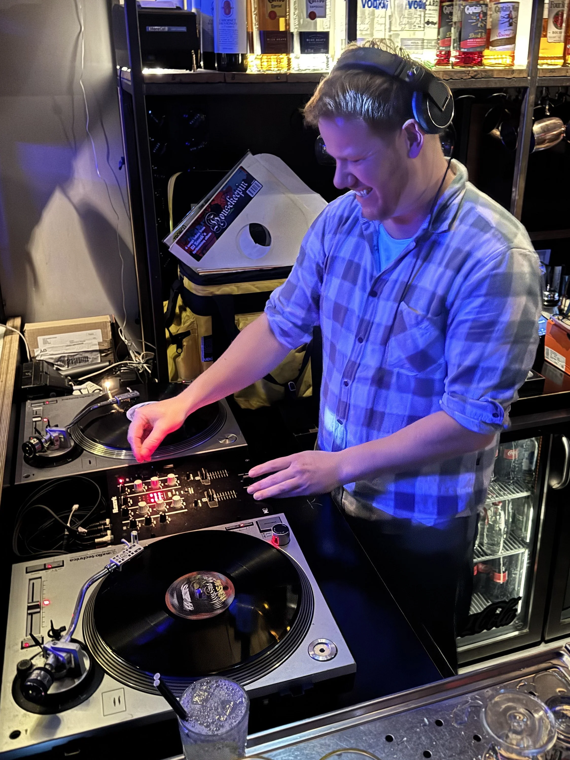 A young man DJing at a bar, wearing headphones, smiling, and operating turntables and a mixer, with bottles of alcohol on the shelves behind him.