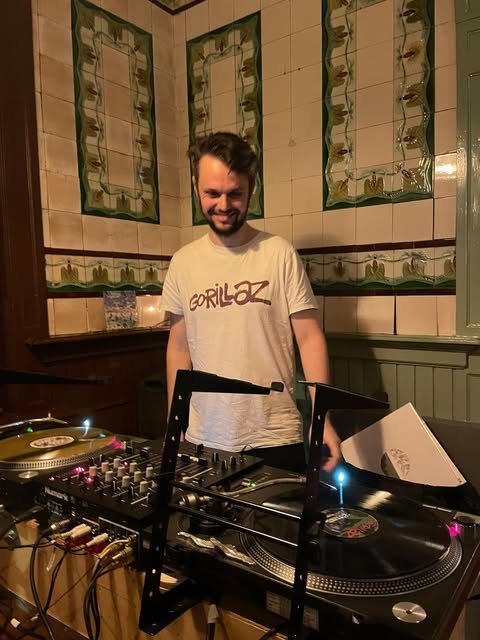 A man with a beard and dark hair, smiling, standing behind DJ equipment in a decorated indoor space with green and beige tile walls.