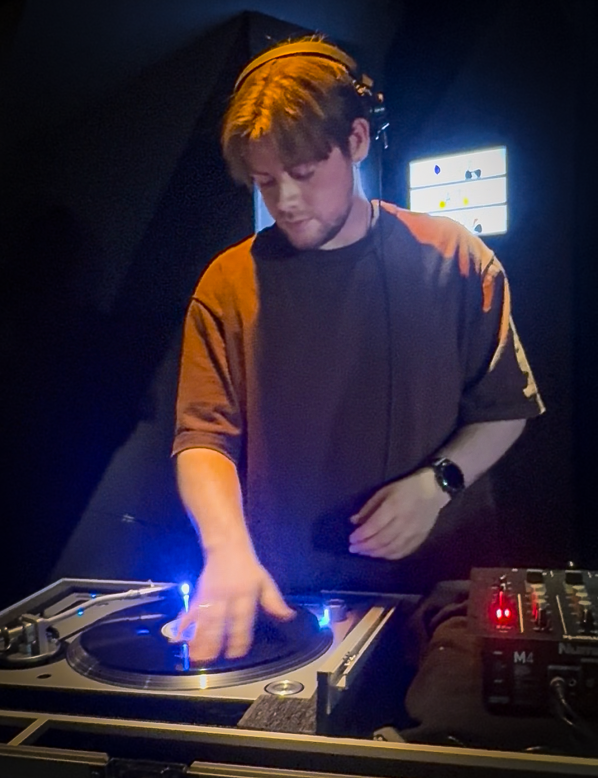 A male DJ standing behind a turntable, adjusting a record with one hand, wearing a dark shirt and a watch, in a dark environment with a bright screen in the background.