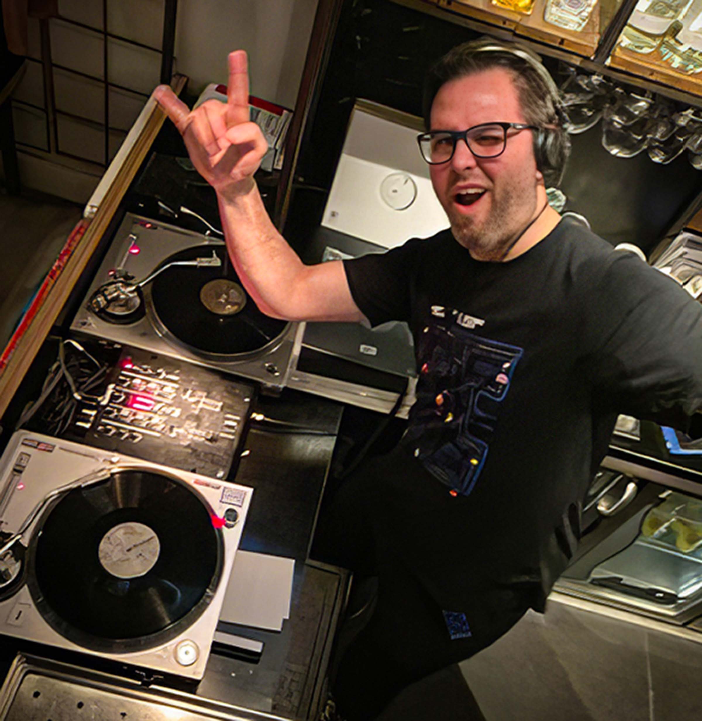 A person with glasses and curly hair making a rock hand gesture while standing next to DJ equipment in a record store or cafe.