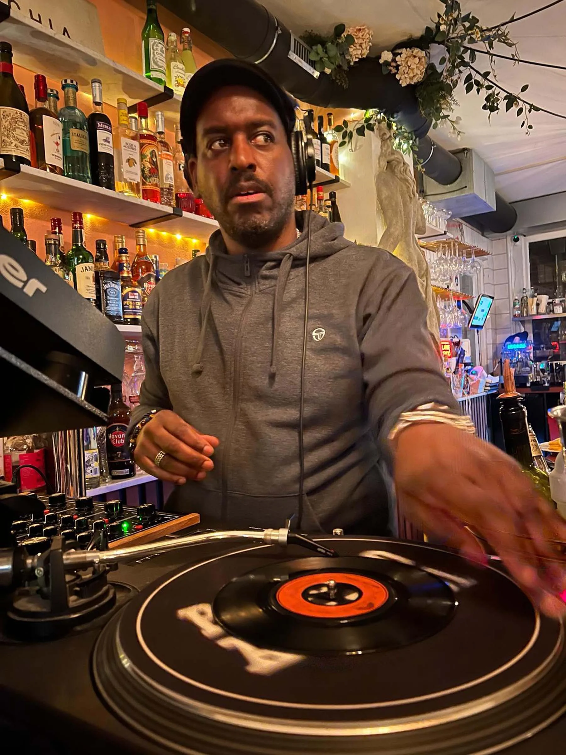 A man wearing headphones DJing at a bar or nightclub, with a vinyl record on a turntable in front of him. Shelves with liquor bottles and decorative plants are visible behind him.