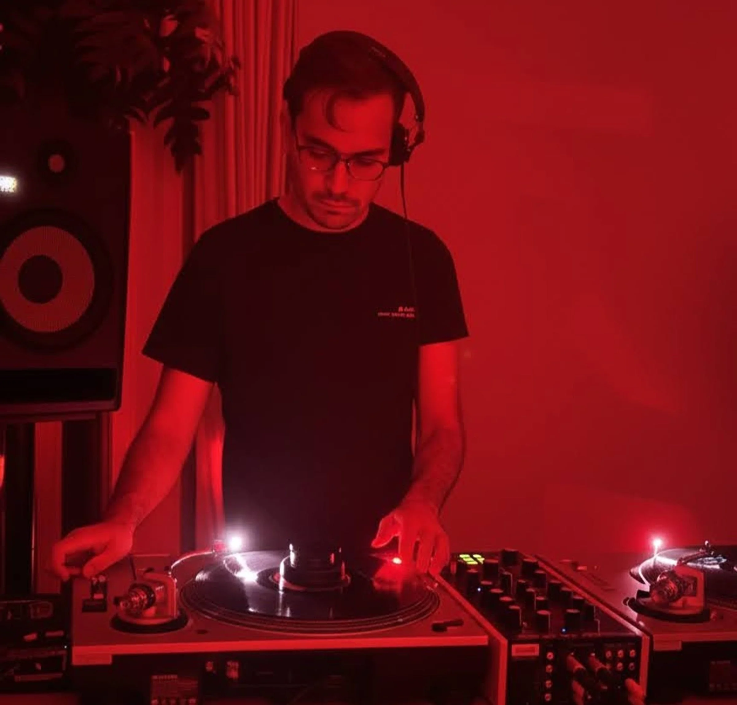 A person wearing headphones and glasses operating a DJ turntable against a red-lit background.