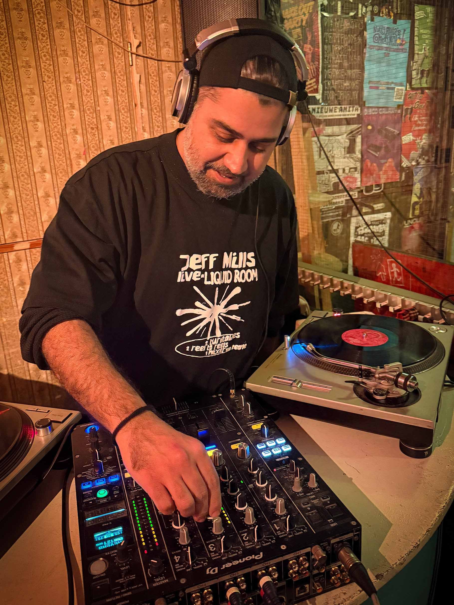 A DJ with gray facial hair wearing a black cap backwards and a black T-shirt playing music on a DJ mixer and turntable in a vintage-style room with wooden walls and colorful posters.