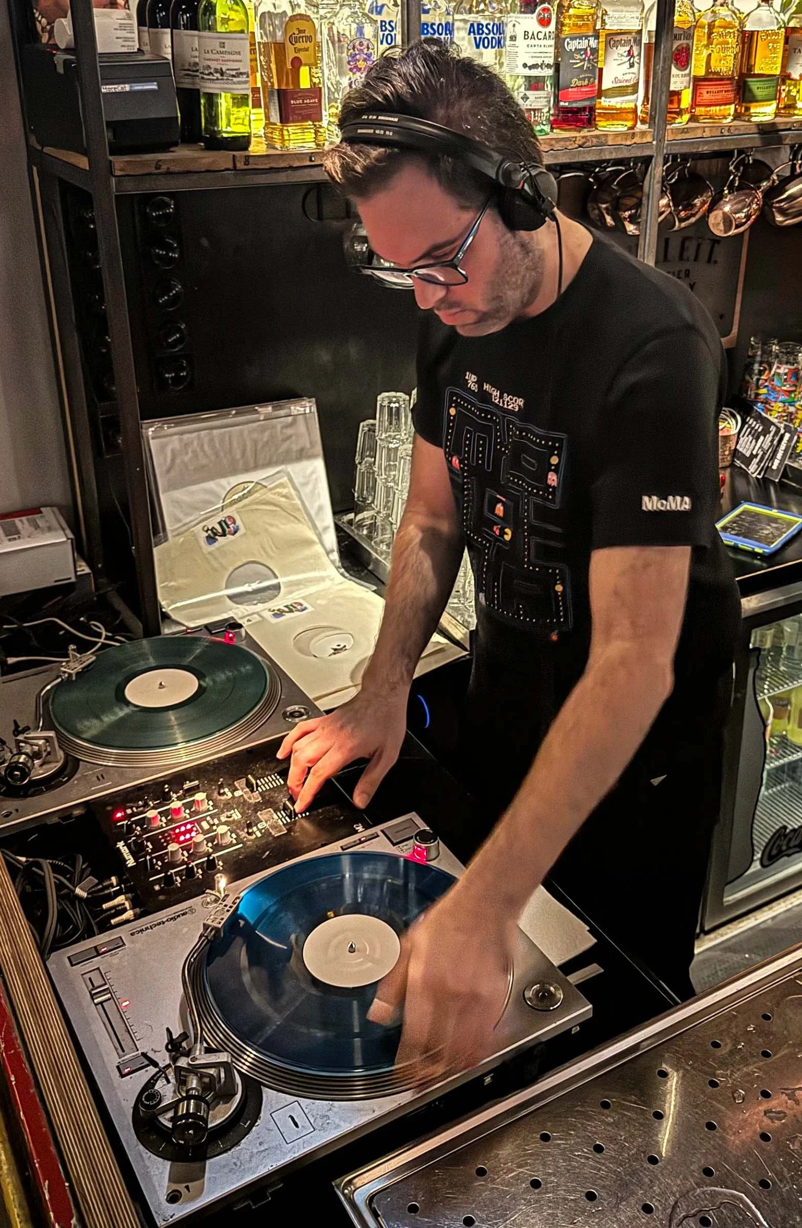 A man with glasses and headphones DJing with two turntables and a mixer, decorated with liquor bottles on shelves in the background.