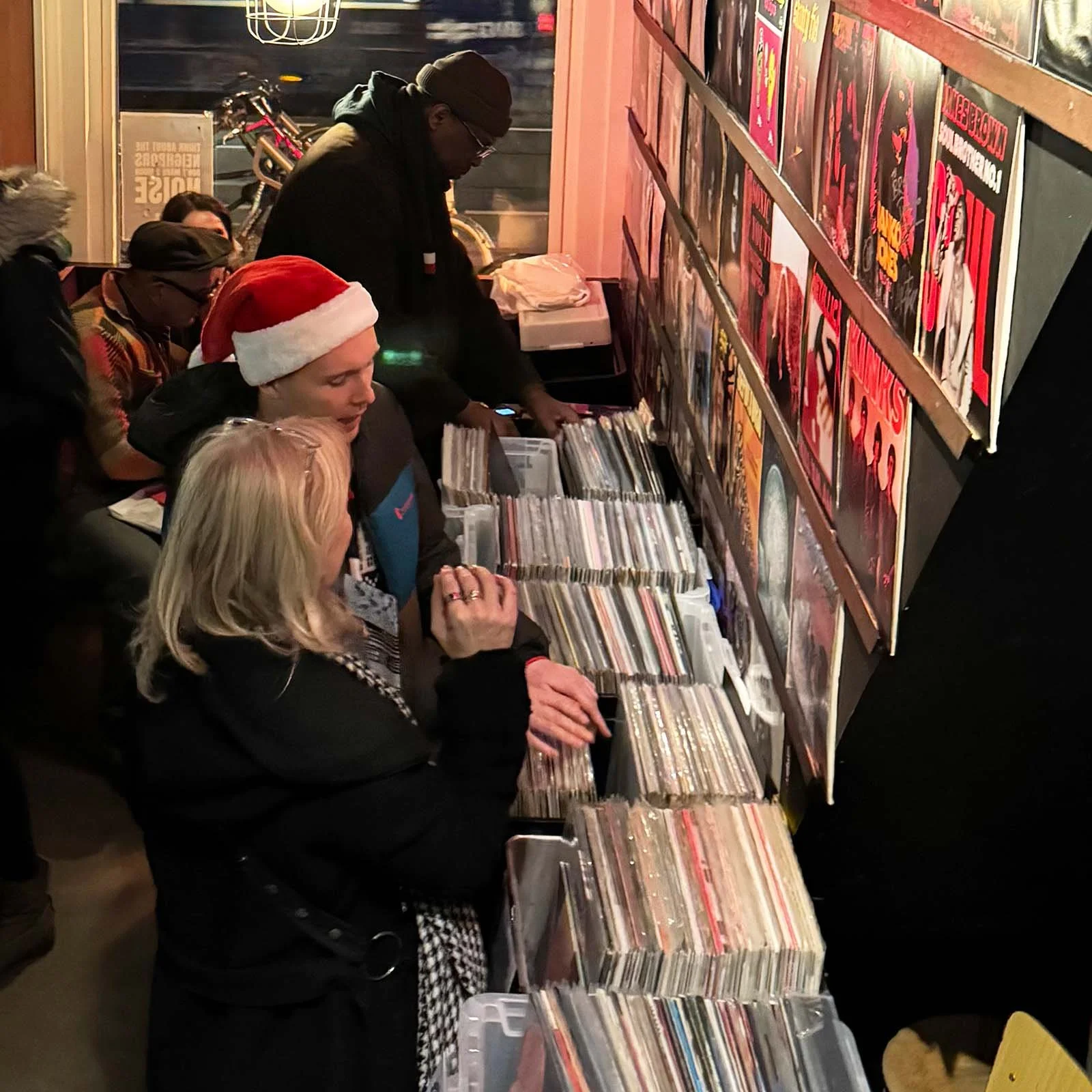 People shopping for vinyl records at an Amsterdam Vinyl Club live event, some wearing holiday hats, with records displayed on tables and artwork on the wall.