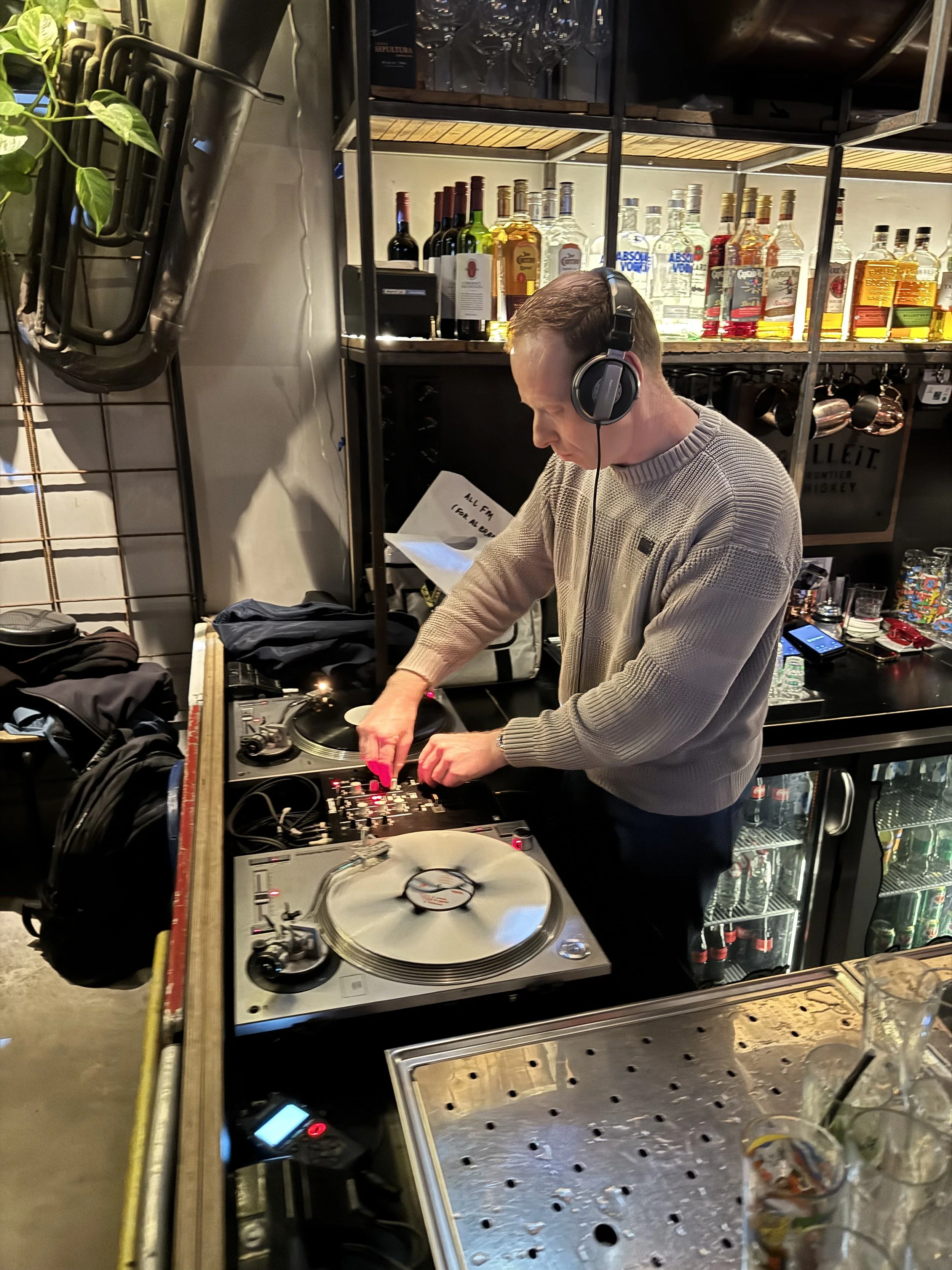 A DJ wearing a gray sweater and headphones, manipulating turntables and mixer in a bar or club setting with alcohol bottles on shelves in the background.