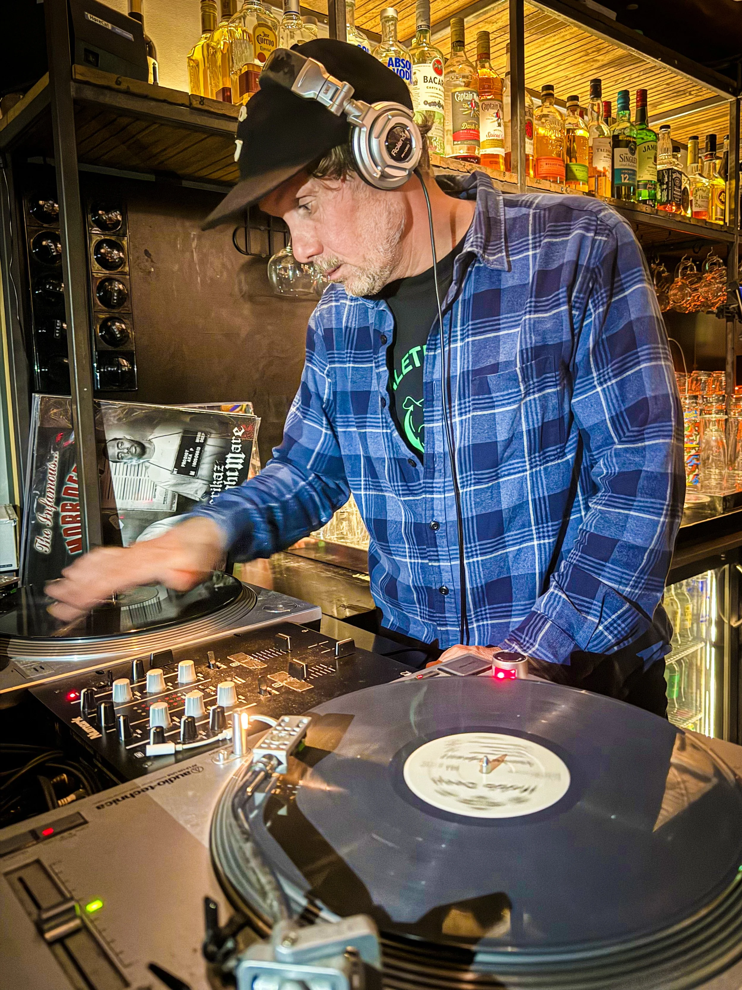 A DJ wearing a black cap backwards, plaid shirt, and headphones is mixing music on a turntable in a bar with bottles of alcohol on shelves in the background.