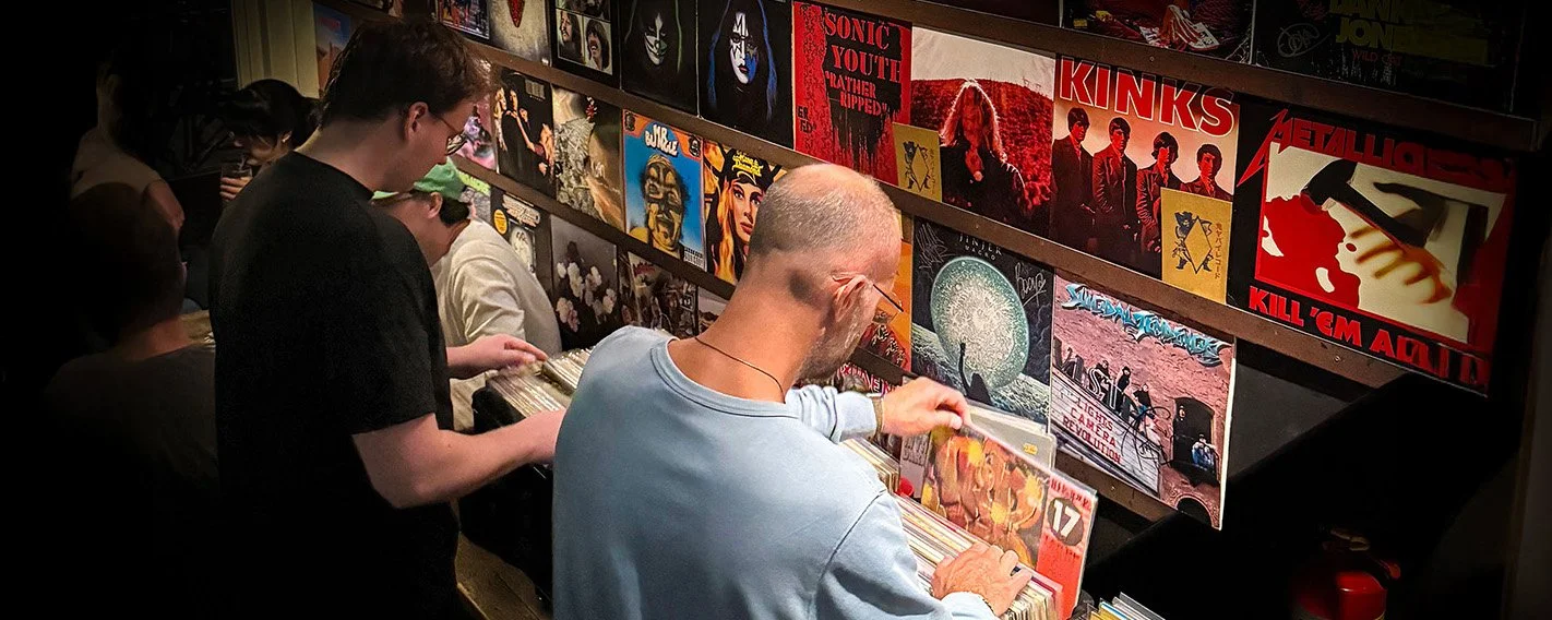 People browsing vinyl records at an Amsterdam Vinyl Club live event. There are album covers displayed on shelves, including bands like Metallica, Kinks, and Sonic Youth.