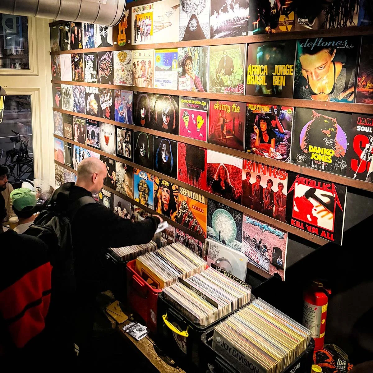 People browsing vinyl records at an Amsterdam Vinyl Club event, with album covers on the wall featuring various artists and bands.