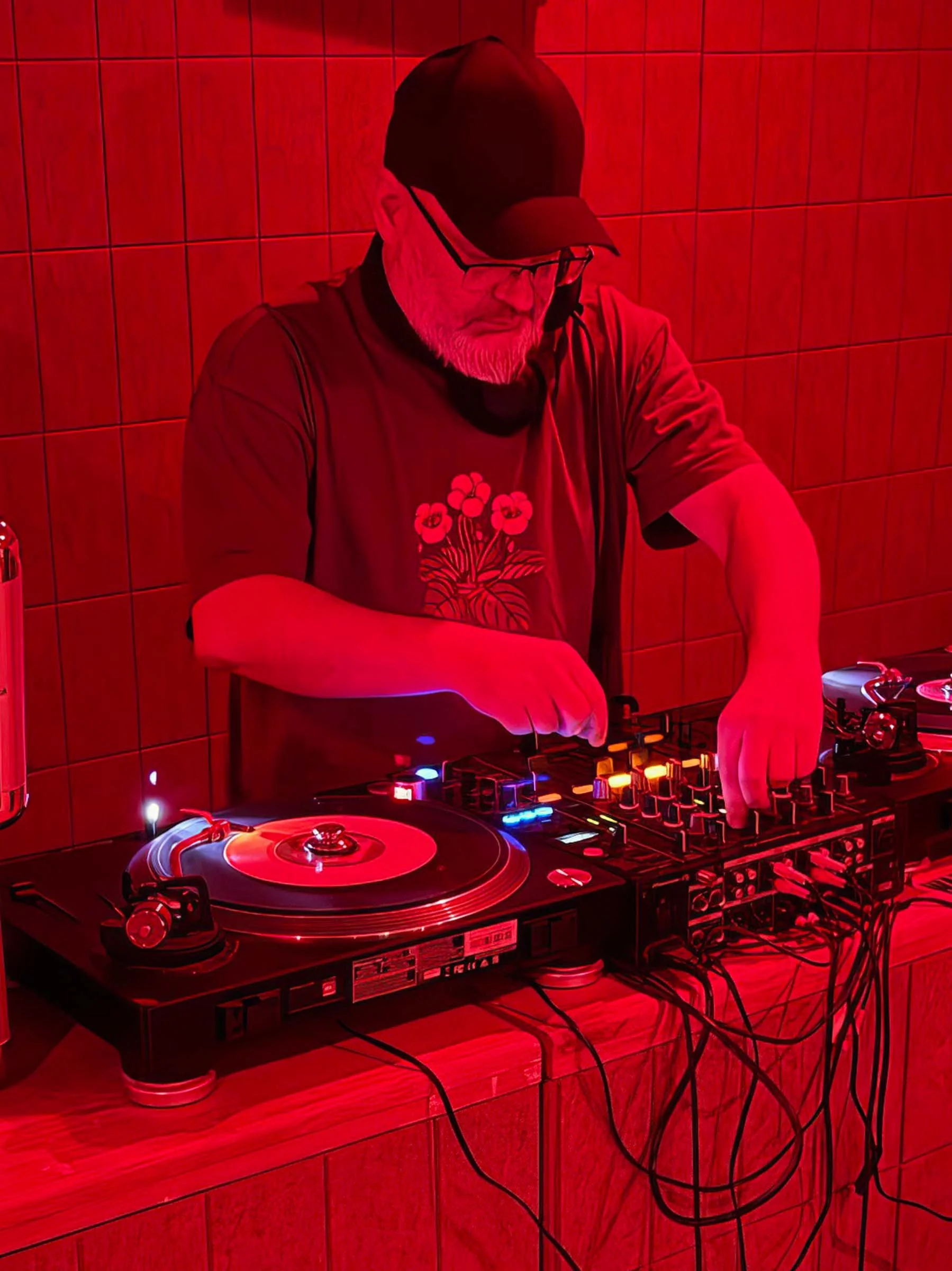 A DJ wearing a black cap and glasses is mixing music on turntables and a DJ mixer, illuminated by red lighting against a tiled wall background.