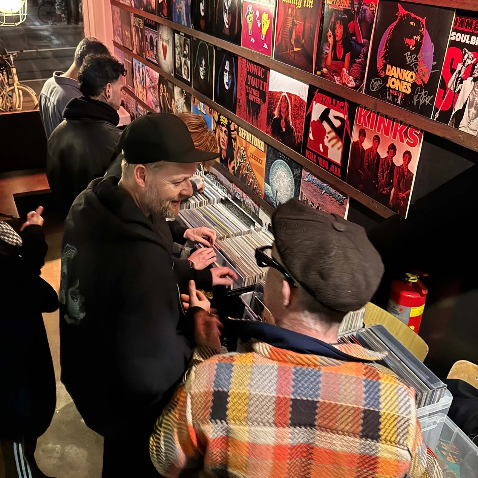 People browsing vinyl records at an Amsterdam Vinyl Club live event, with colorful album covers displayed on the wall.