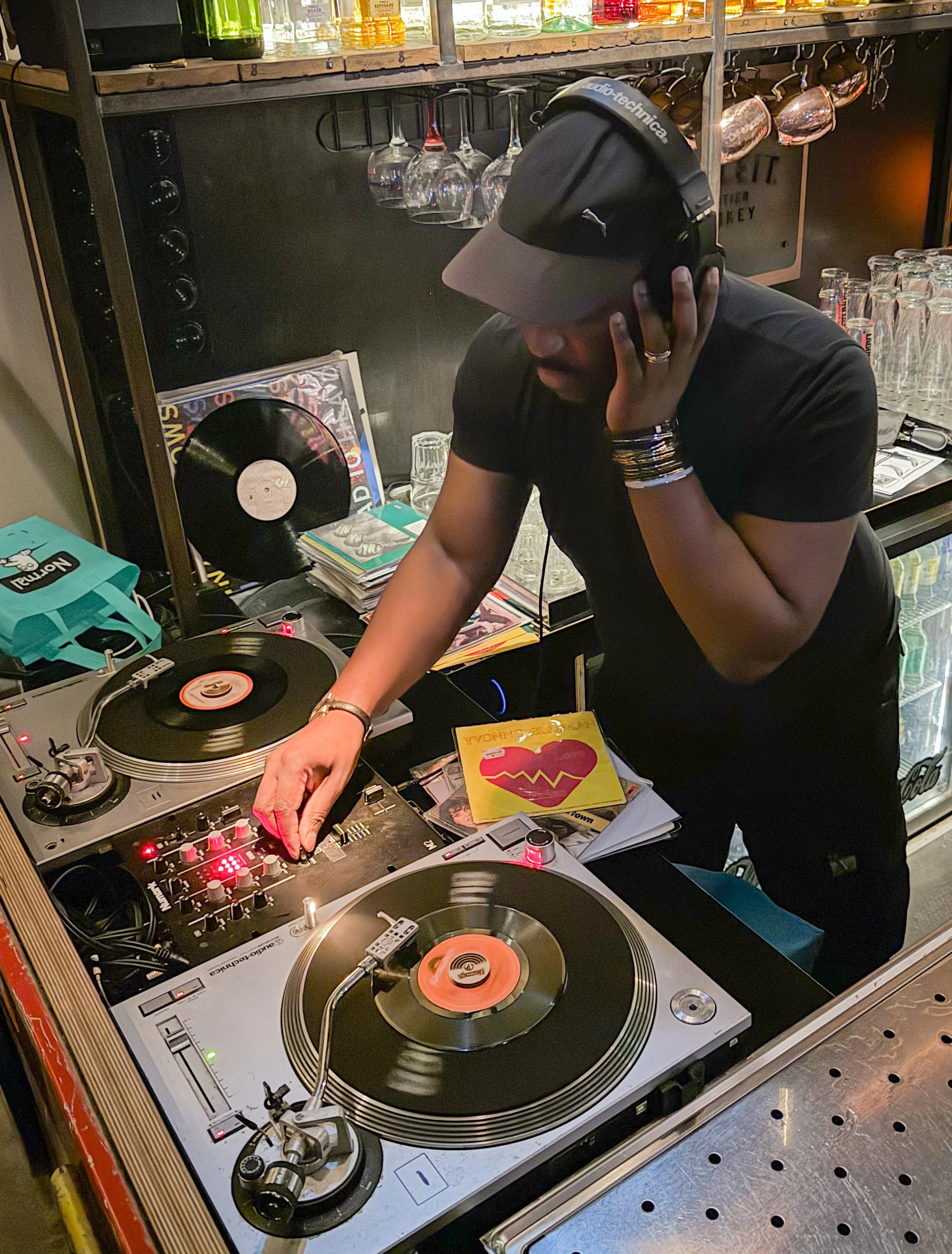 A DJ is performing at a bar, wearing a black cap, black shirt, and headphones, with one hand on a turntable and the other adjusting the headphone on his ear. The setup includes two turntables, a mixer, vinyl records, and some music magazines. Behind him are shelves with glasses and some bottles.